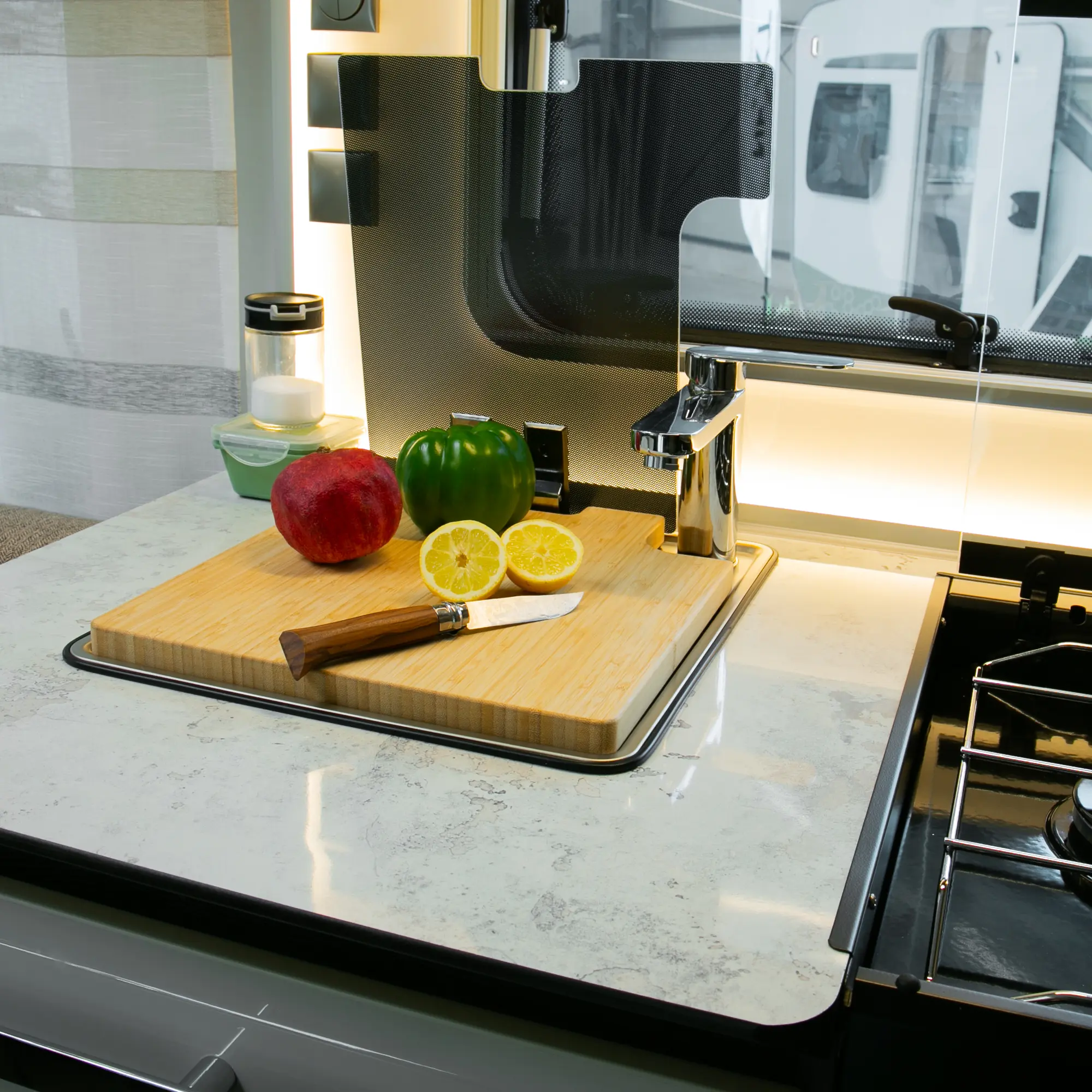 Tabla de cortar y tapa de fregadero your GEAR Burano de bambú en una cocina de caravana, con cuchillo, pimiento verde, limón y manzana sobre la superficie, junto a un grifo y estufa.