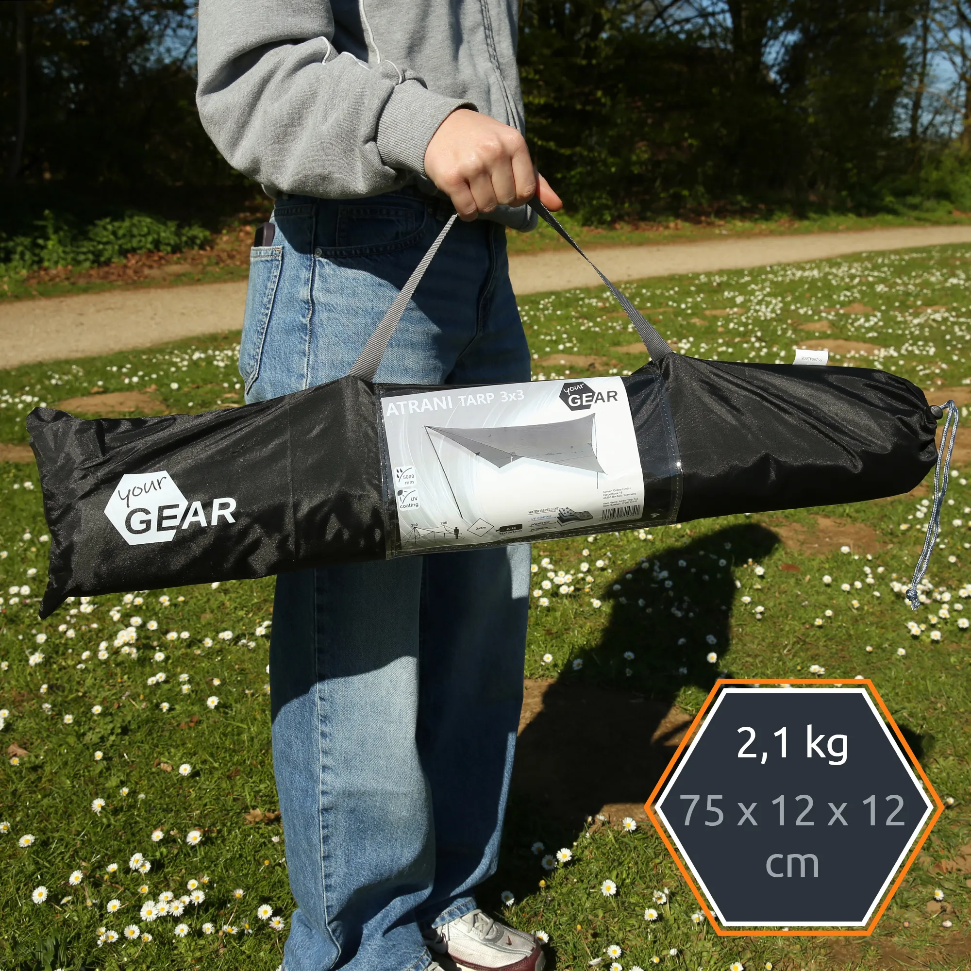 Person holding black carry bag for "your GEAR" Atrani 3x3 m grey waterproof tarp, visible label showing tarp diagram, straps and packed cylindrical storage bag against grassy park background