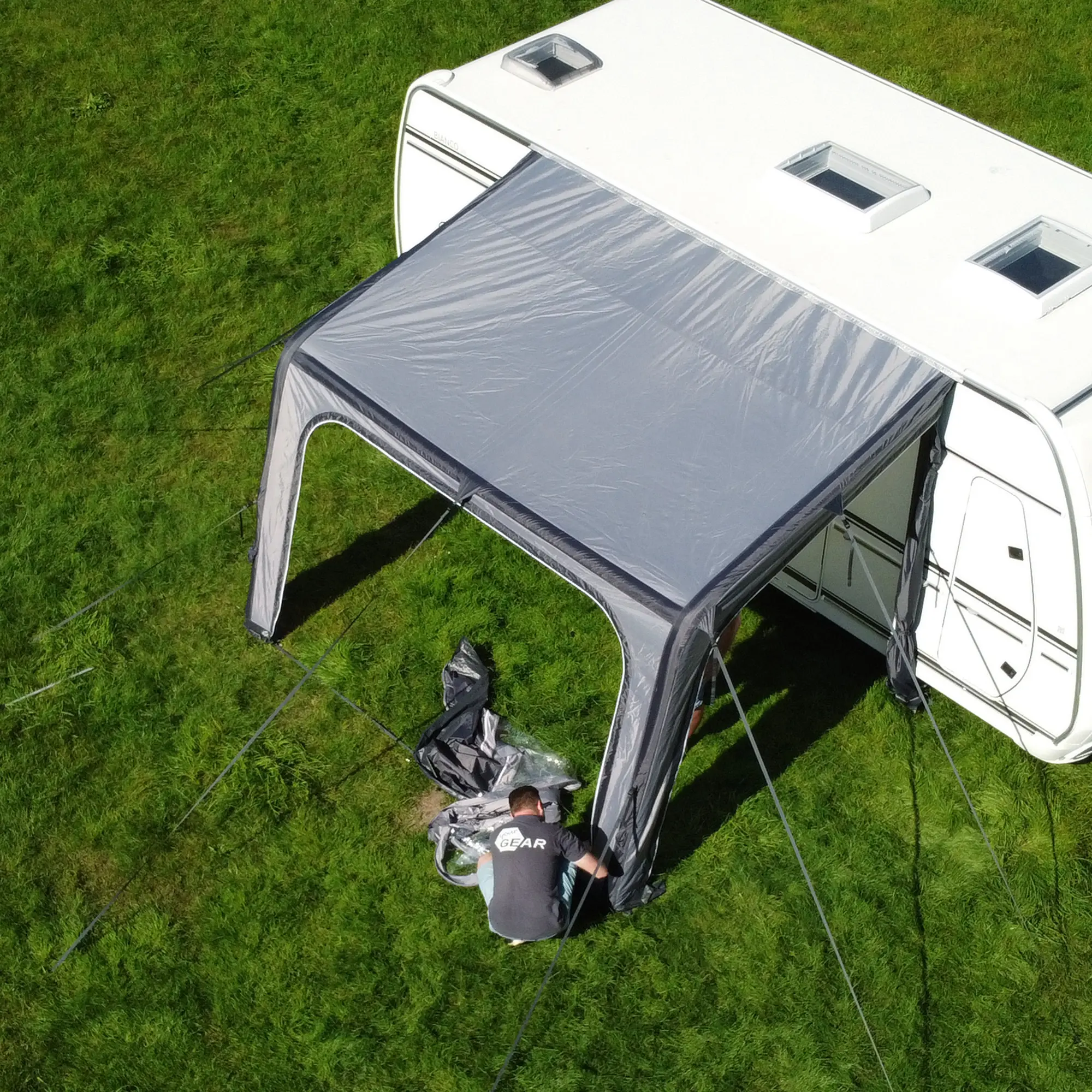 Avance de un hombre montando el toldo inflable your GEAR Porto 340 Air gris en una caravana blanca, mostrando su estructura ligera, resistente y fácil instalación sobre césped verde. Ideal para protección solar y lluvia.