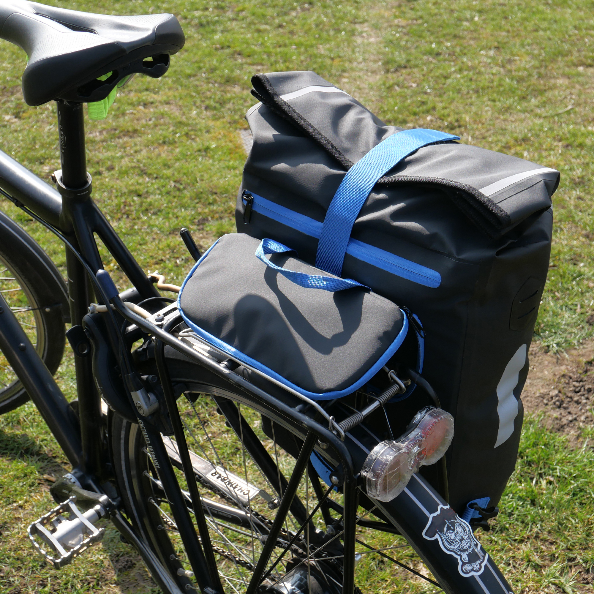 yourGEAR Arco 19L 3-in-1 waterproof bike pannier, rucksack, and laptop bag in black with blue accents, attached to a bicycle rear rack, featuring a roll-top closure and reflective elements.