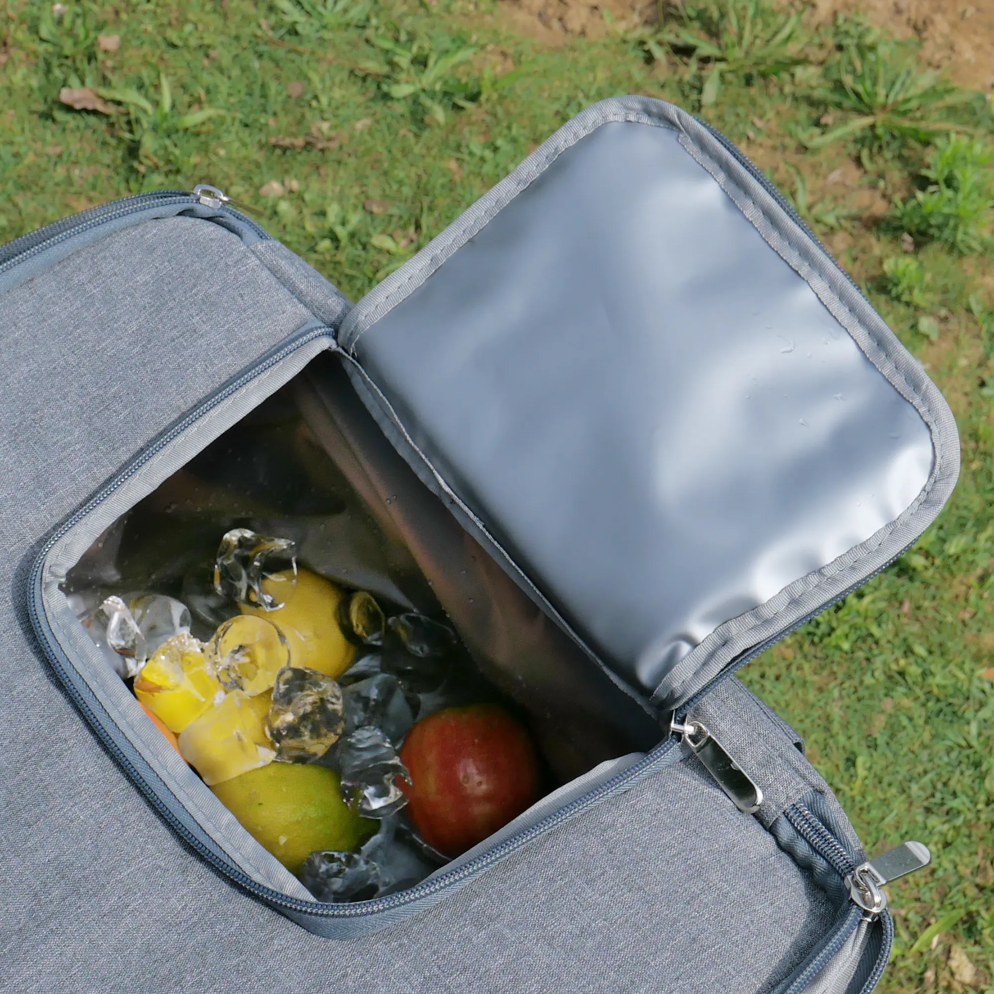 your GEAR foldable cooler bag Cavoli, 30 liter insulated bag in grey, open top with zipper, visible PEVA lining, filled with fruits and ice cubes, shown outdoors on grass.