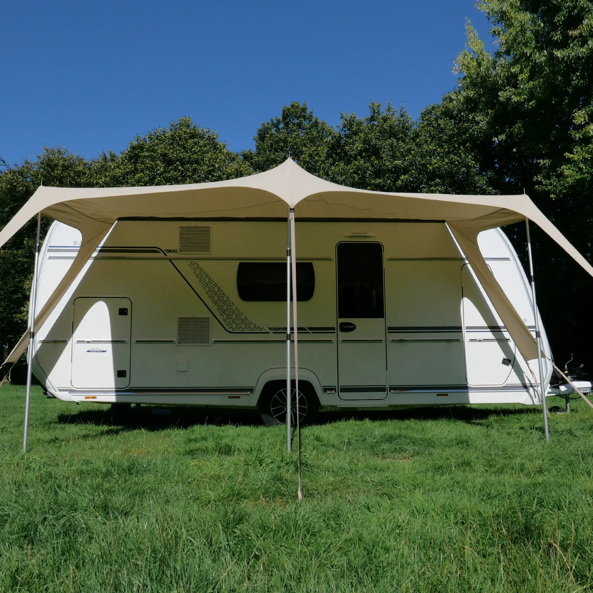 Beiges your GEAR Scilla TC Caravan Tarp Sonnensegel 400x250 cm als Sonnendach am Wohnwagen montiert, bietet großzügigen Schatten und UV-Schutz, steht auf vier Stangen auf einer Wiese, Bäume im Hintergrund.