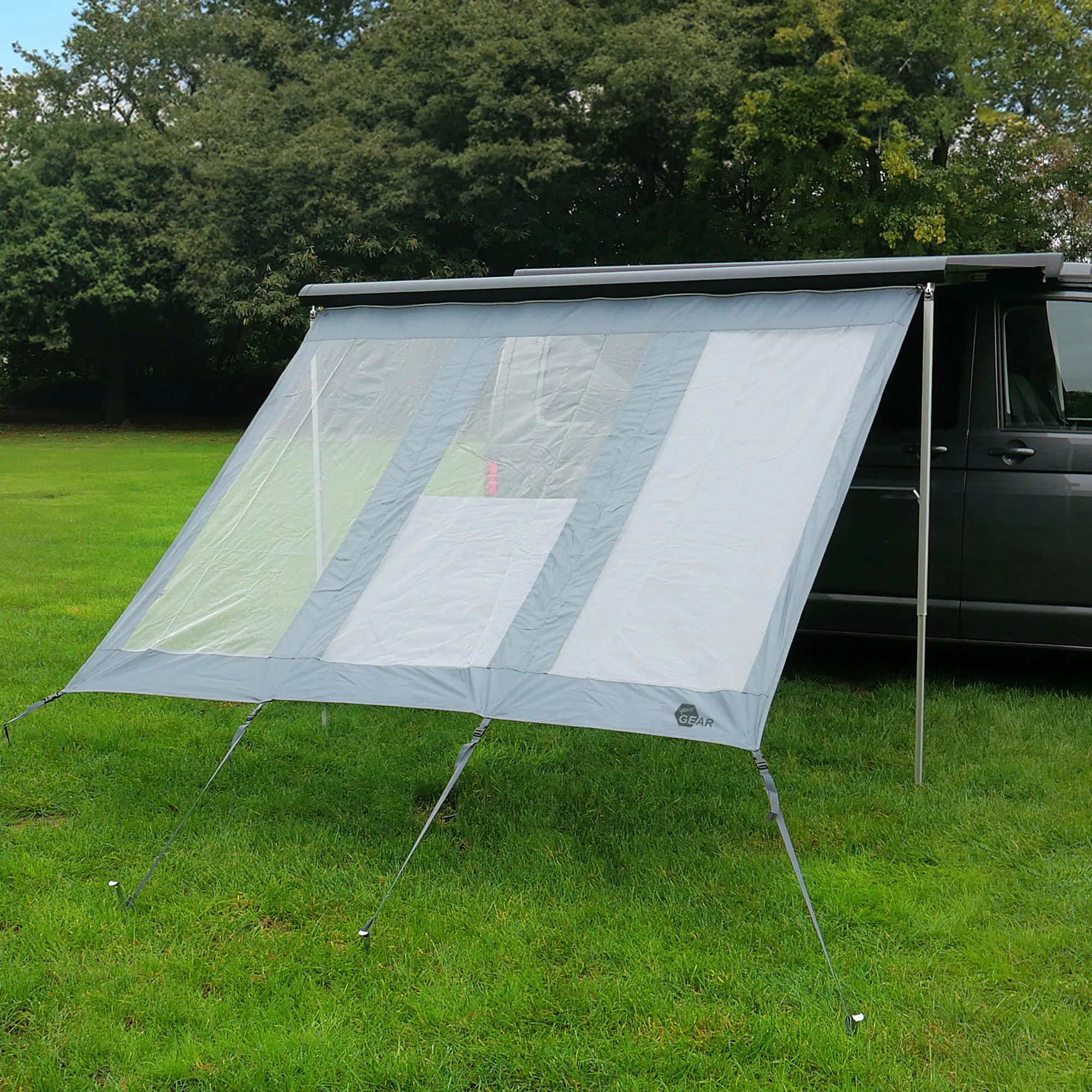 your GEAR Panorama Wall 2,4 x 2 m als Markisen-Vorderwand am Fahrzeug montiert, grau mit transparenten Einsätzen, schützt vor Sonne, Wind und Regen, auf einer Wiese vor Bäumen aufgestellt.