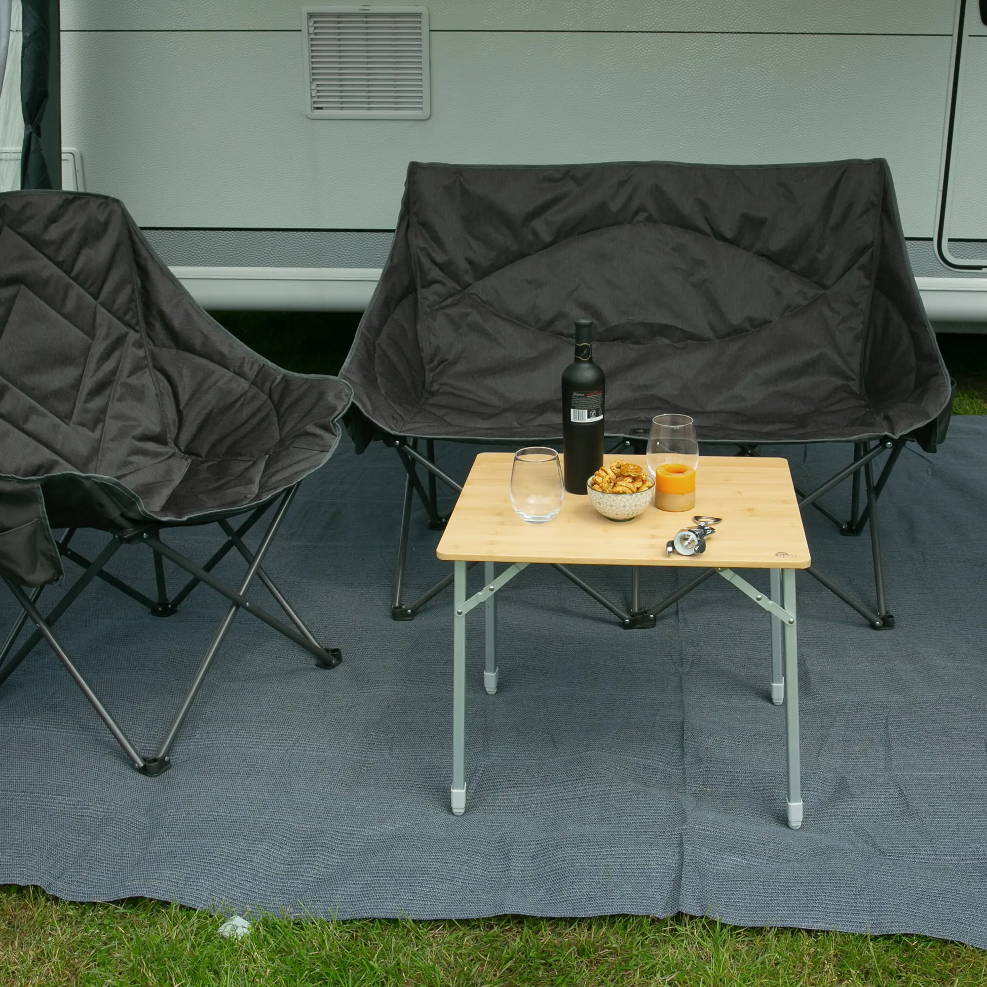 Bambus Campingtisch your GEAR Portofino M mit höhenverstellbaren Aluminiumbeinen, rechteckiger Tischplatte, aufgestellt zwischen zwei Campingstühlen, belegt mit Getränken und Snacks, Outdoor-Einsatz vor Wohnmobil.