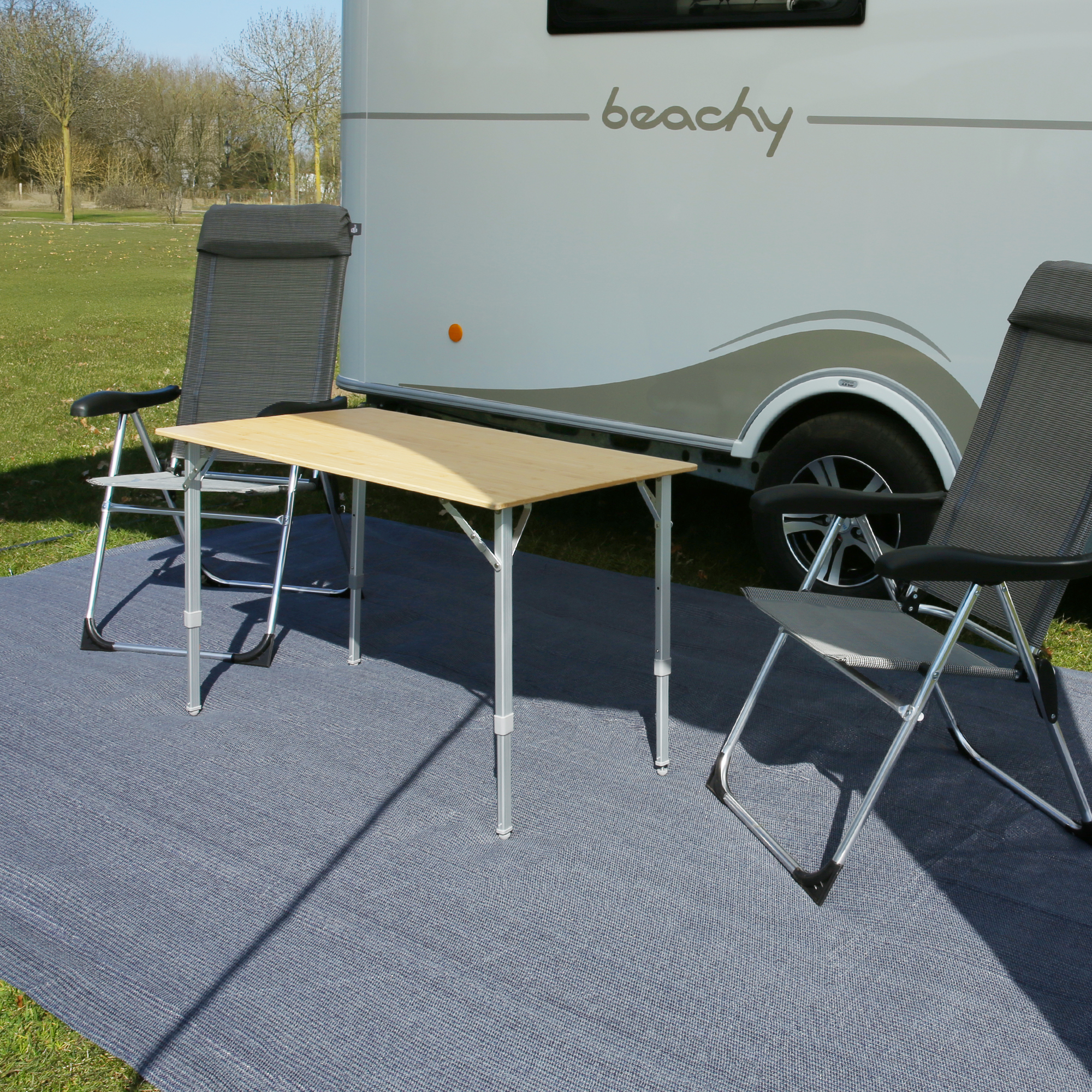 Dos sillas de camping your GEAR Novara con respaldo alto, estructura de aluminio, reposacabezas acolchado y tejido gris resistente, colocadas junto a una mesa exterior frente a una caravana.
