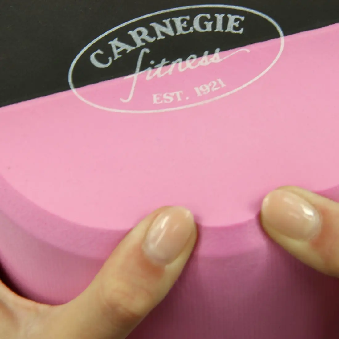Rulo de pilates Carnegie EVA rosa de 90x15 cm, superficie lisa, logo “Carnegie Fitness” impreso en blanco en un extremo, dos manos sujetando la espuma, ideal para yoga, fitness y automasaje.