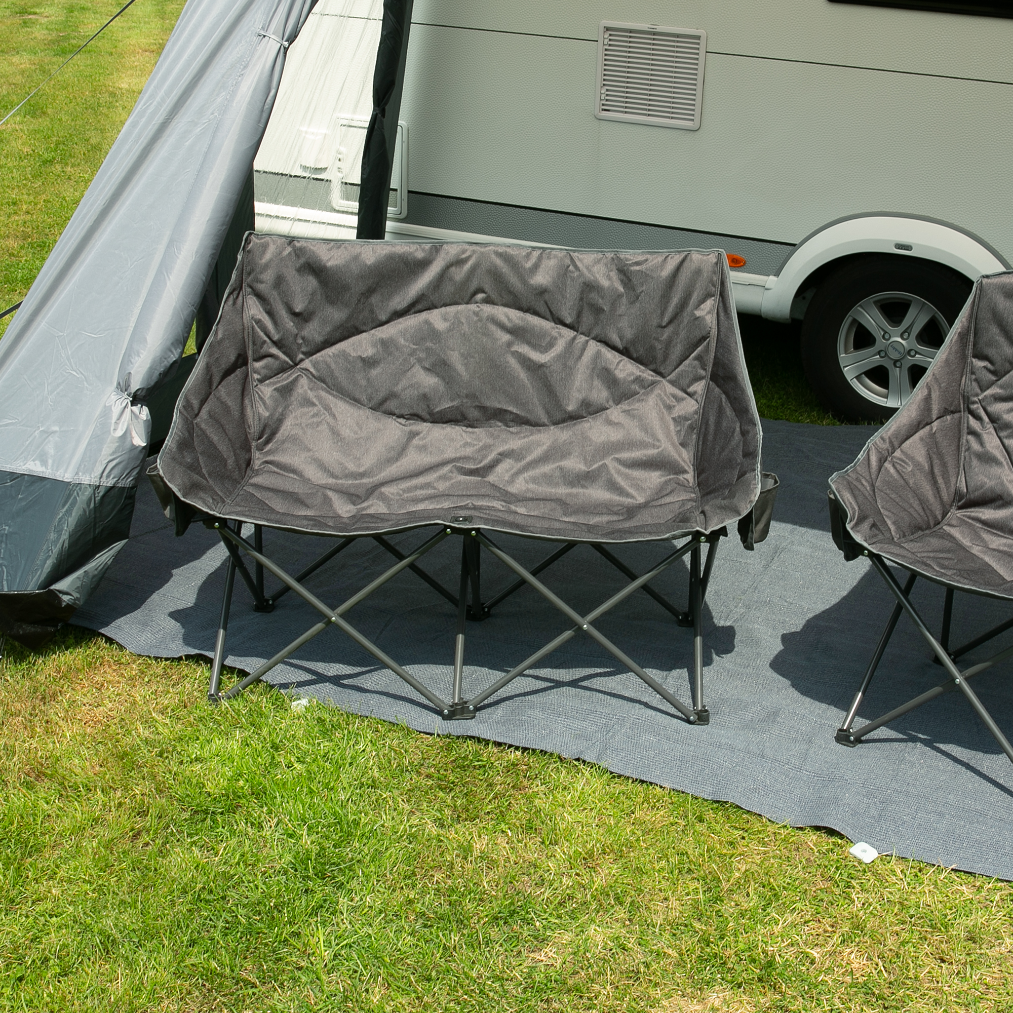 Gepolstertes your GEAR Campingsofa Modena für 2 Personen, XXL-Größe, extra breite Sitzfläche, robustes Stahlgestell, grau, faltbar, steht vor Wohnwagen auf Campingplatz. Outdoor-Campingstuhl, hohe Belastbarkeit.