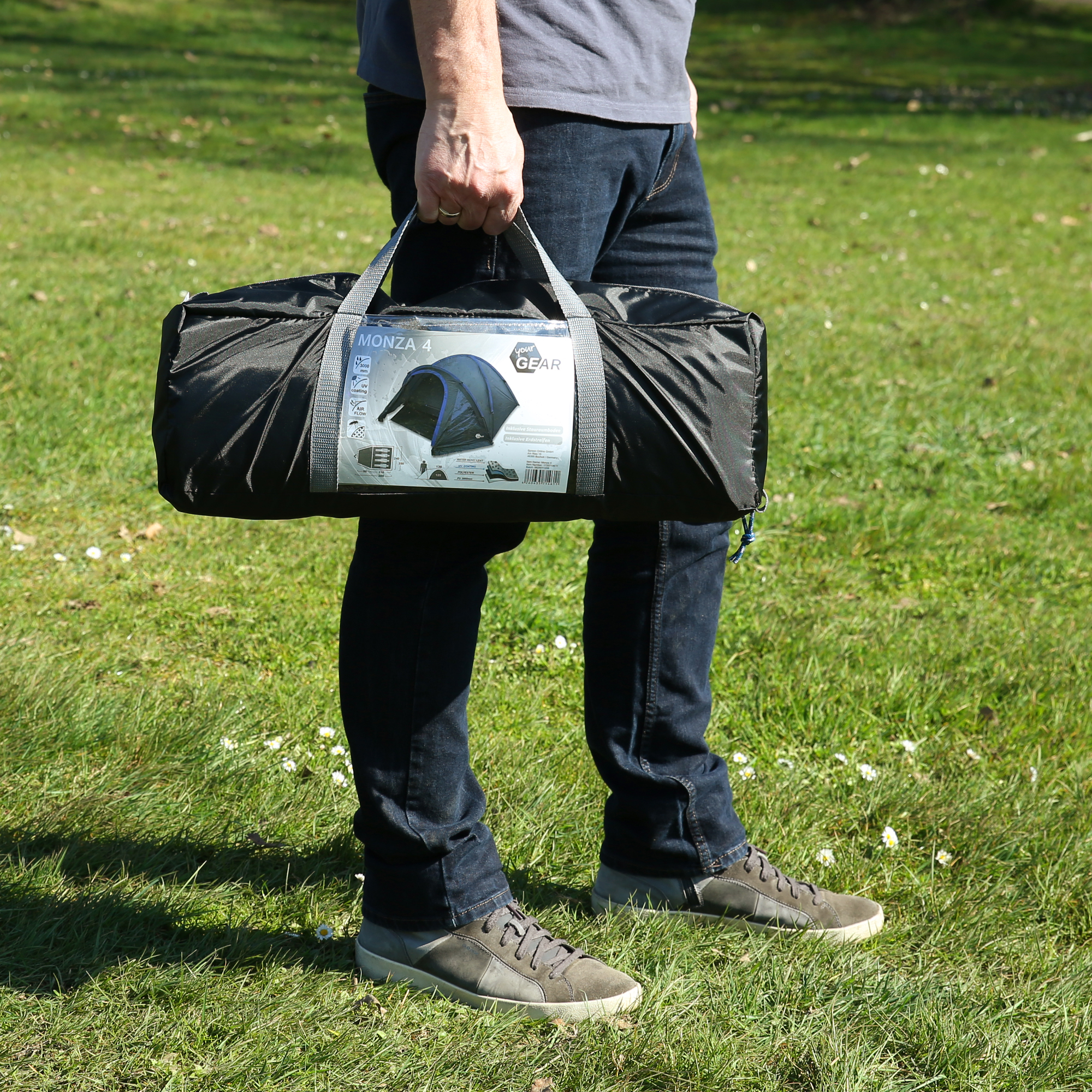 Persona sosteniendo una bolsa de transporte negra del your GEAR Zelt Monza 4 personas, con etiqueta visible mostrando imagen del producto y detalles como Kuppelzelt, ventana, UV50+ y 3000 mm, en césped.