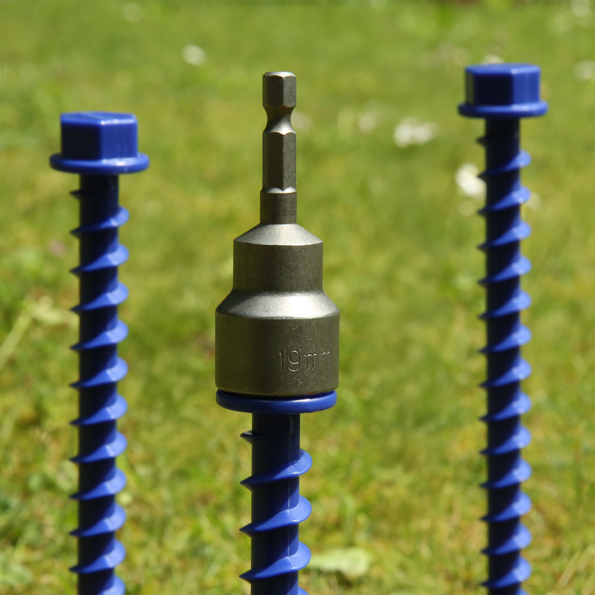 Primo piano di un attacco esagonale metallico 19mm montato su un chiodo a vite corto blu per tenda/caravan, spirale evidente e cappuccio esagonale blu, sfondo prato sfocato.