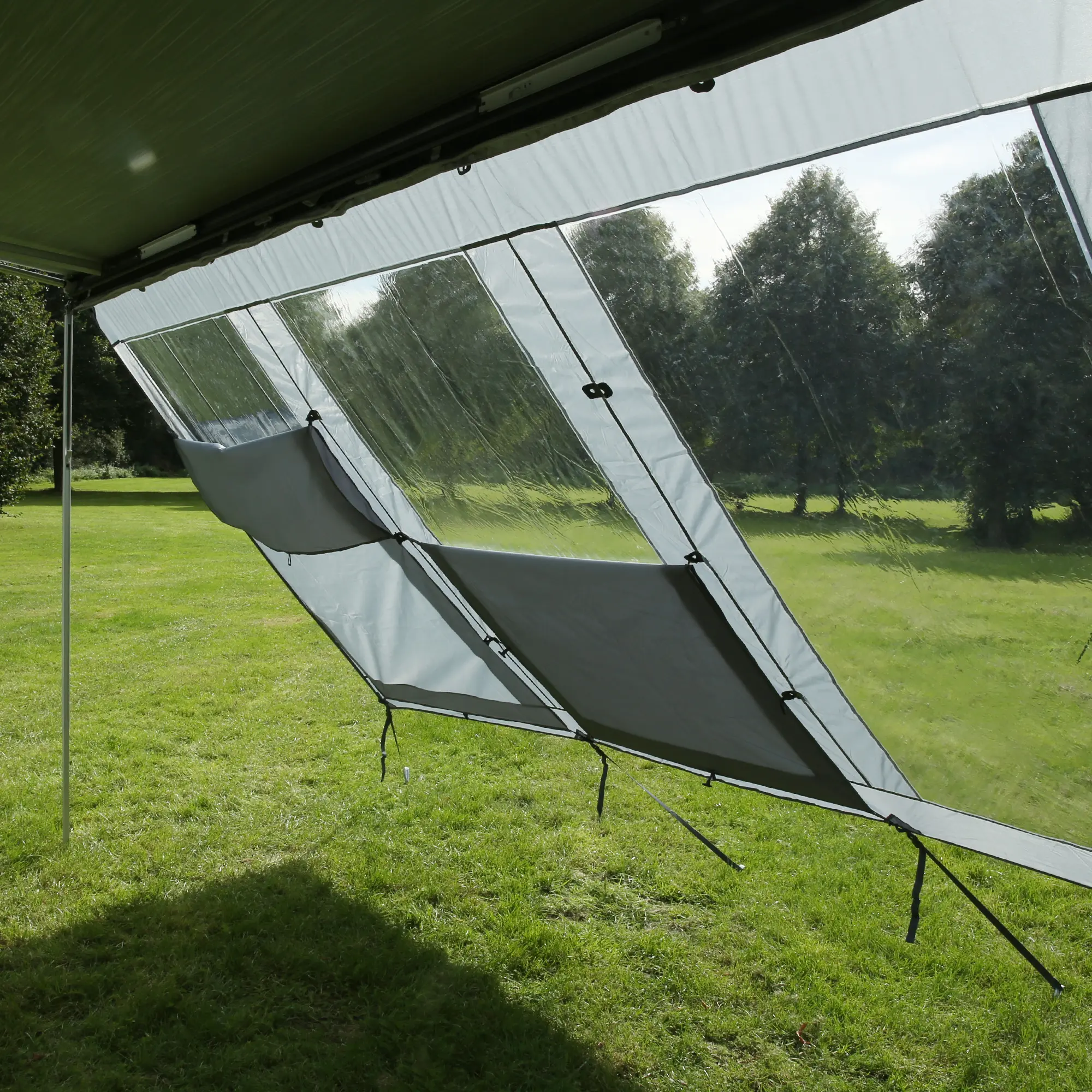 your GEAR Panorama Wall 2,8 x 2 m als transparente Markisenwand mit grauen Sichtschutzflächen, gespannt unter einer Markise auf einer Wiese, schützt vor Sonne, Wind und Regen, ideal für Camping.