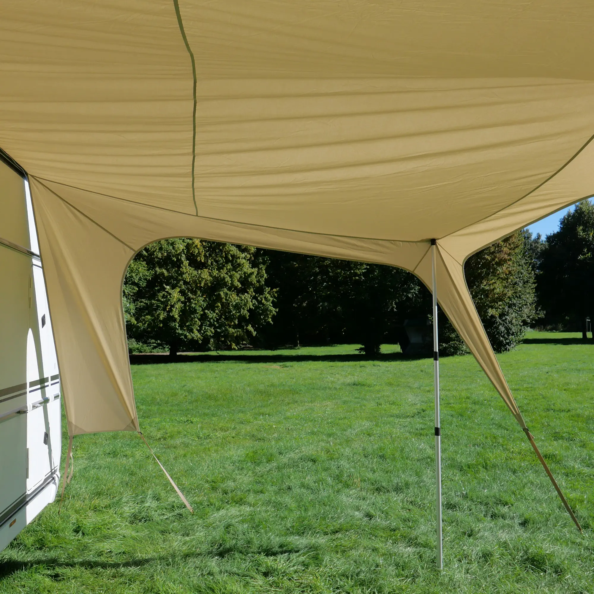 Beiges your GEAR Scilla TC Caravan Tarp Sonnensegel 400x250 cm, montiert am Wohnwagen, gespannt mit Stangen und Abspannseilen, bietet Schatten und UV-Schutz auf einer grünen Wiese.