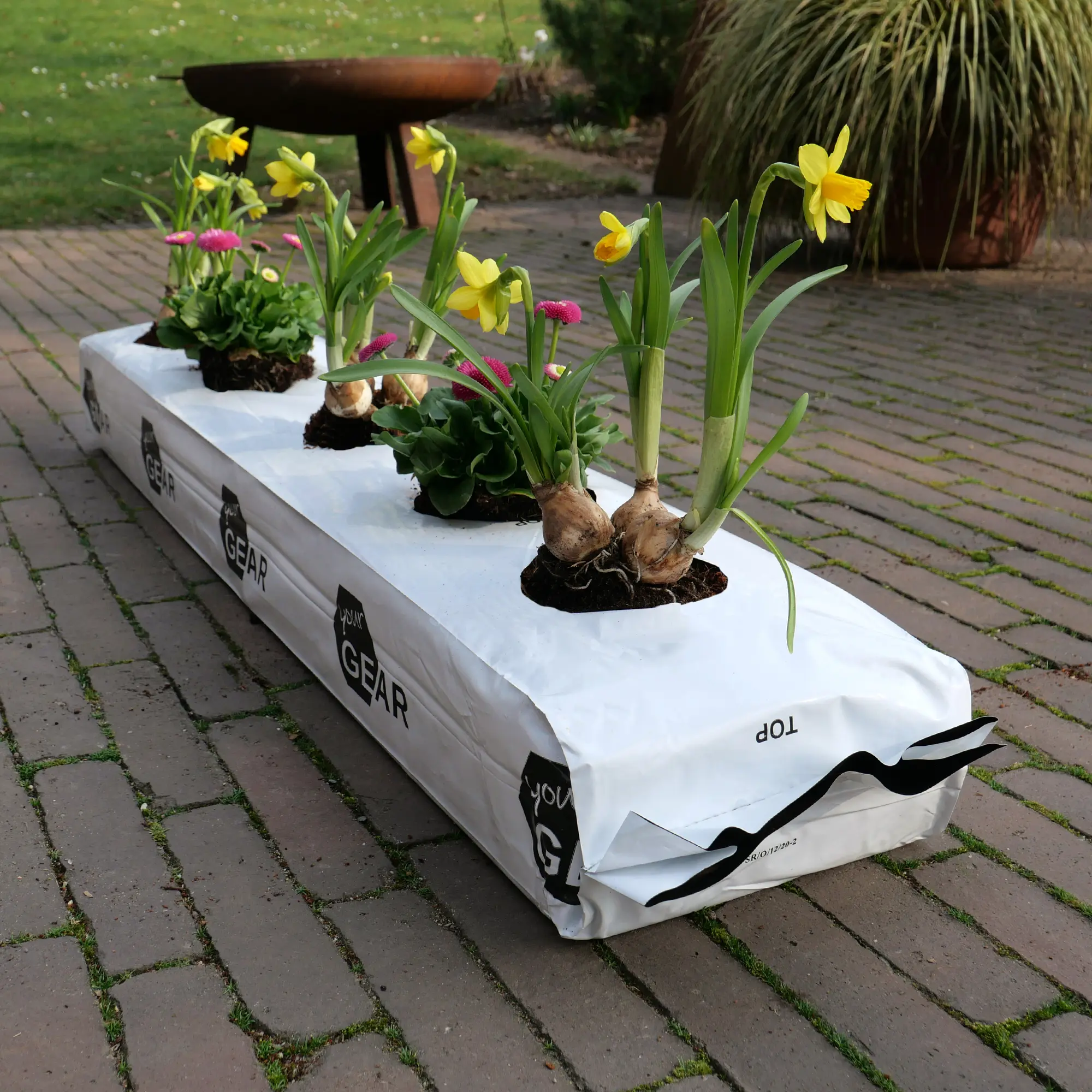 Sacco rettangolare yourGEAR 3x25L Coco Grow Bag bianco con logo, riempito di fibra di cocco pressata, utilizzato come substrato per piante da giardino con fiori gialli e rosa, posizionato su pavimentazione esterna.
