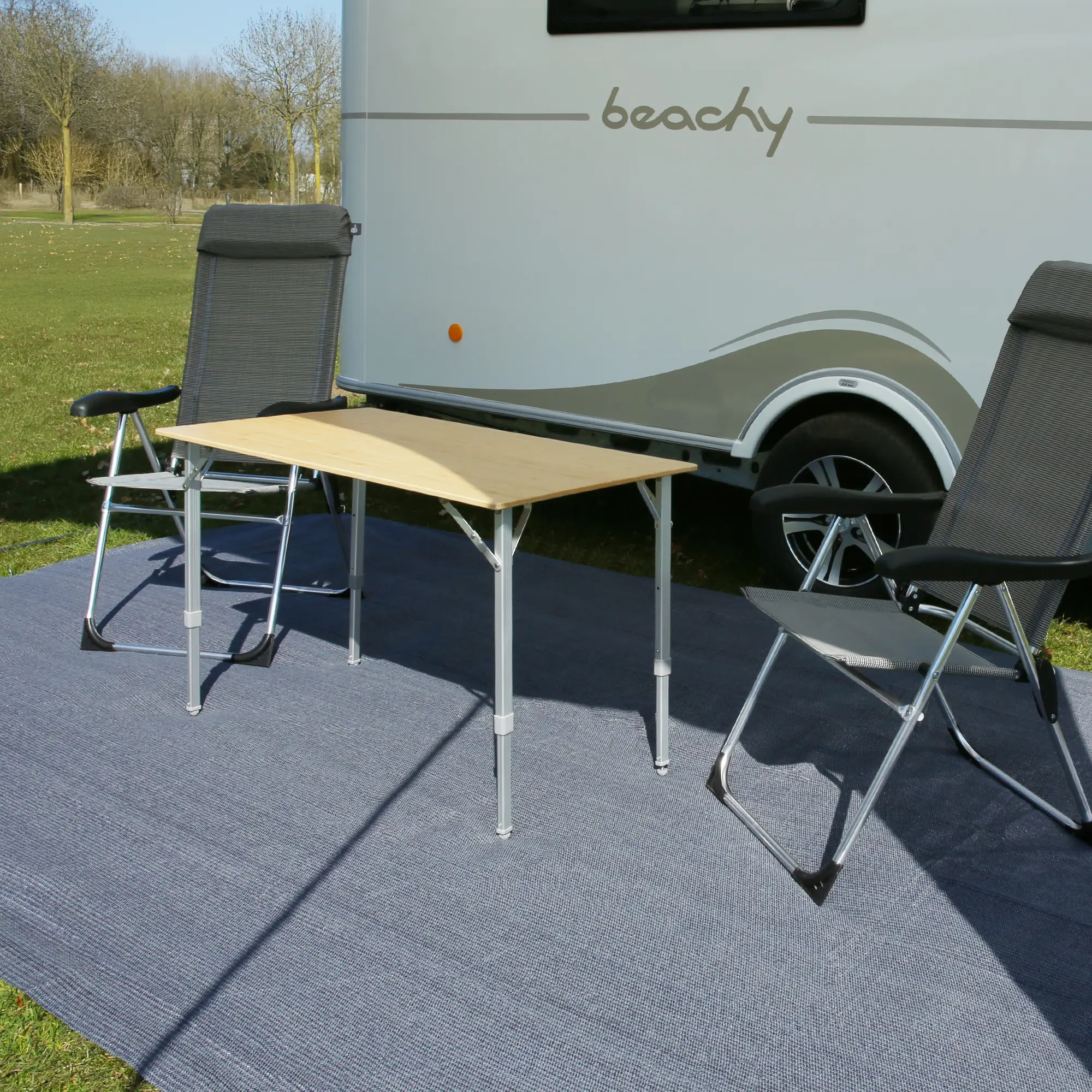 your GEAR Toskana 250 x 300 cm HDPE awning carpet in grey-blue, laid out in front of a caravan with two camping chairs and a table on top, showing its large, durable and weather-resistant surface for outdoor use.