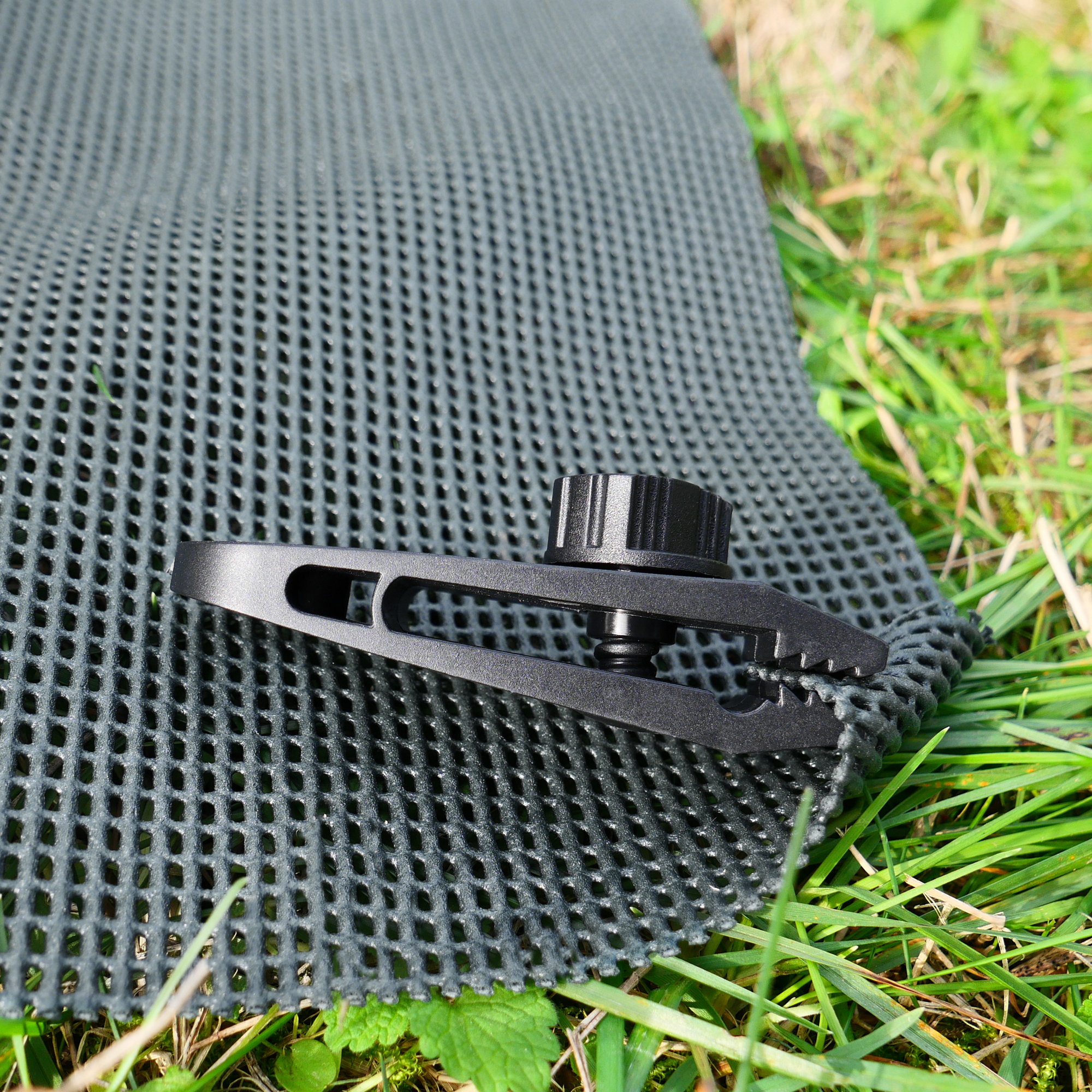 Tapis de sol your GEAR Outdoor Soft 250 x 300 cm, structure à mailles ouvertes gris foncé, fixé avec une pince noire sur l’herbe, idéal pour auvent, camping-car ou tente.