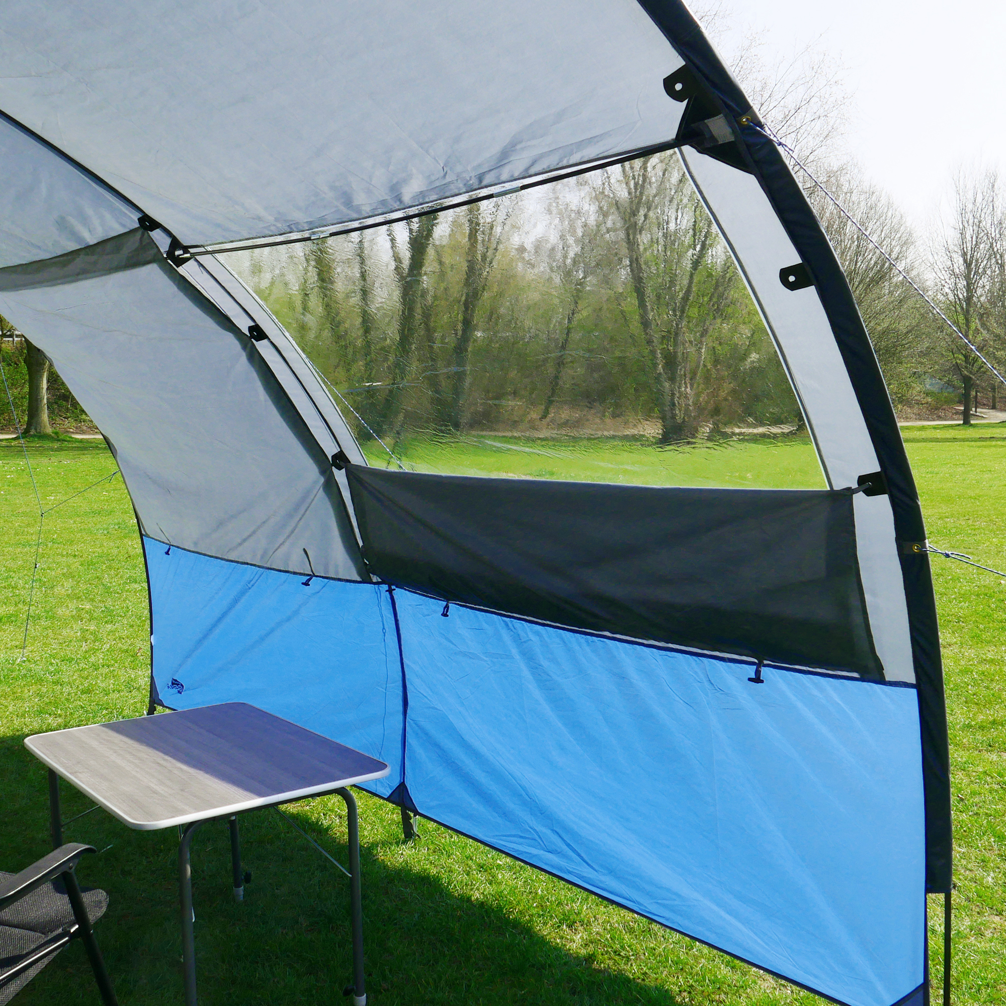 Auvent your GEAR Riviera 350 bleu et gris avec grande fenêtre panoramique, structure légère, protection UV 50+, monté sur pelouse, table de camping noire et grise visible à l’intérieur.