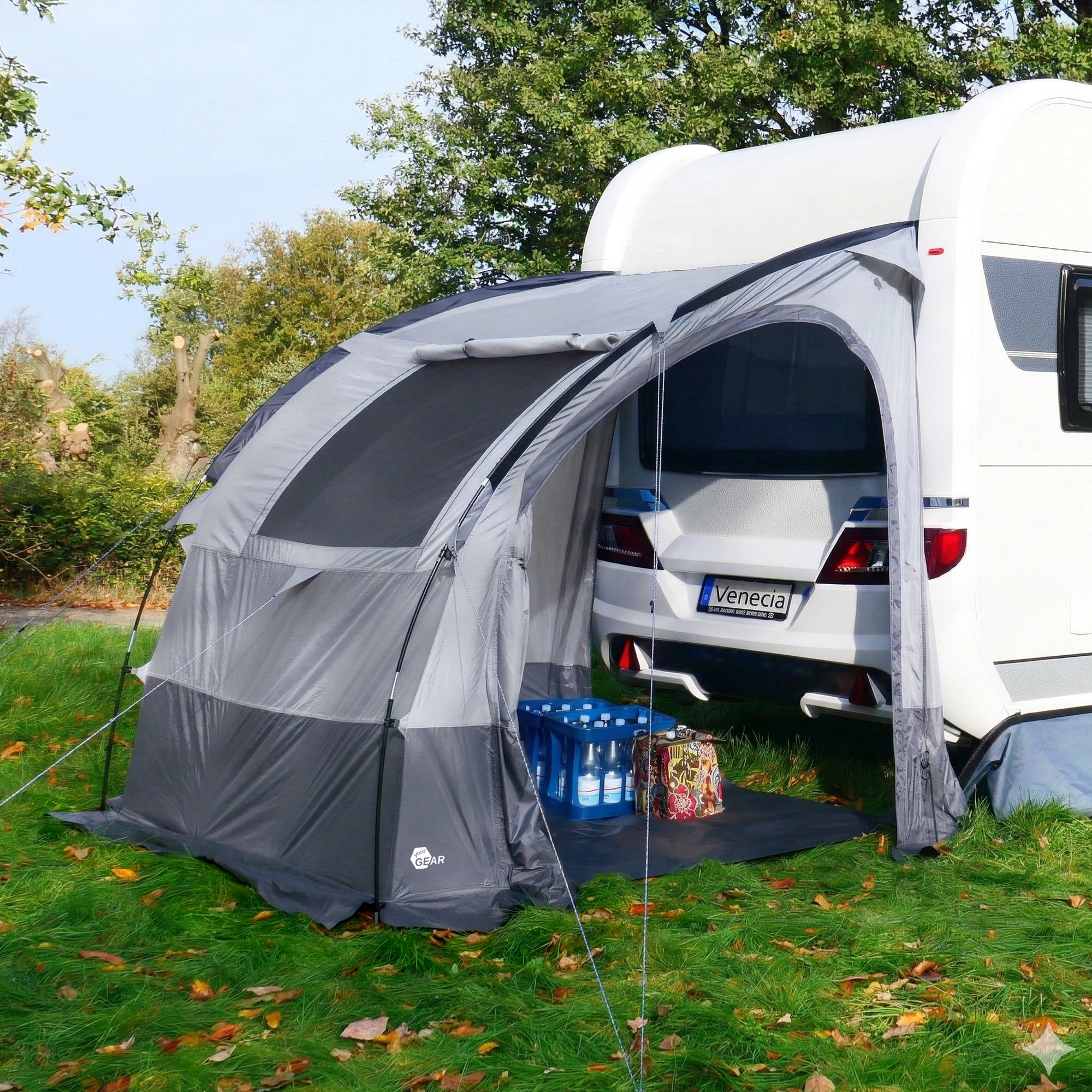 Tente d’extension arrière your GEAR Venecia grise fixée à une caravane, ouverture large en arc, arceaux gonflables ou en fibre, solinigé inclus, utilisée comme abri de rangement/cuisine avec caisse d’eau visible.
