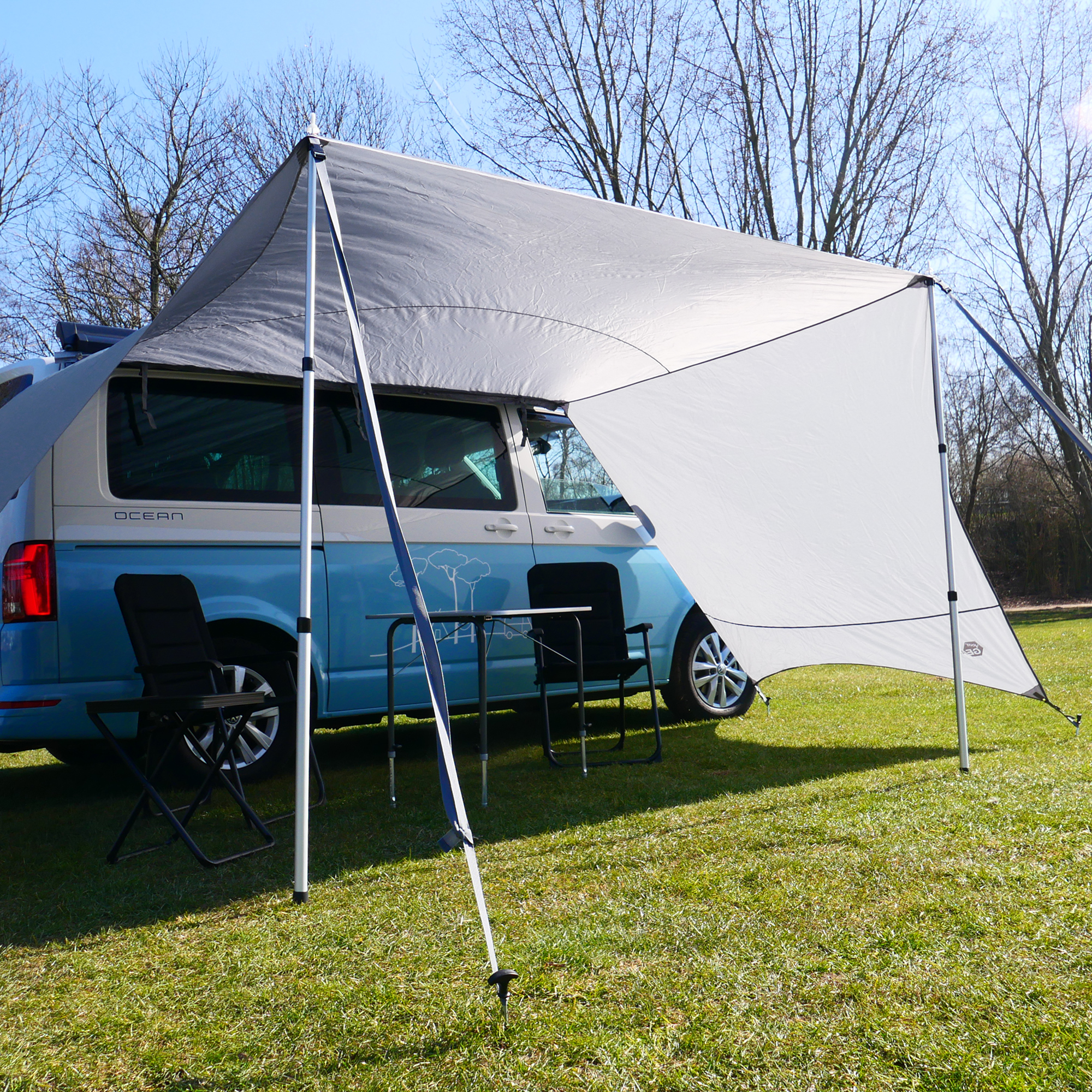 your GEAR Tropea Bus awning UV 50+ with side walls, mounted on a blue camper van; includes 2 aluminum poles, provides sun and rain protection, shown set up on grass with camping chairs and table.