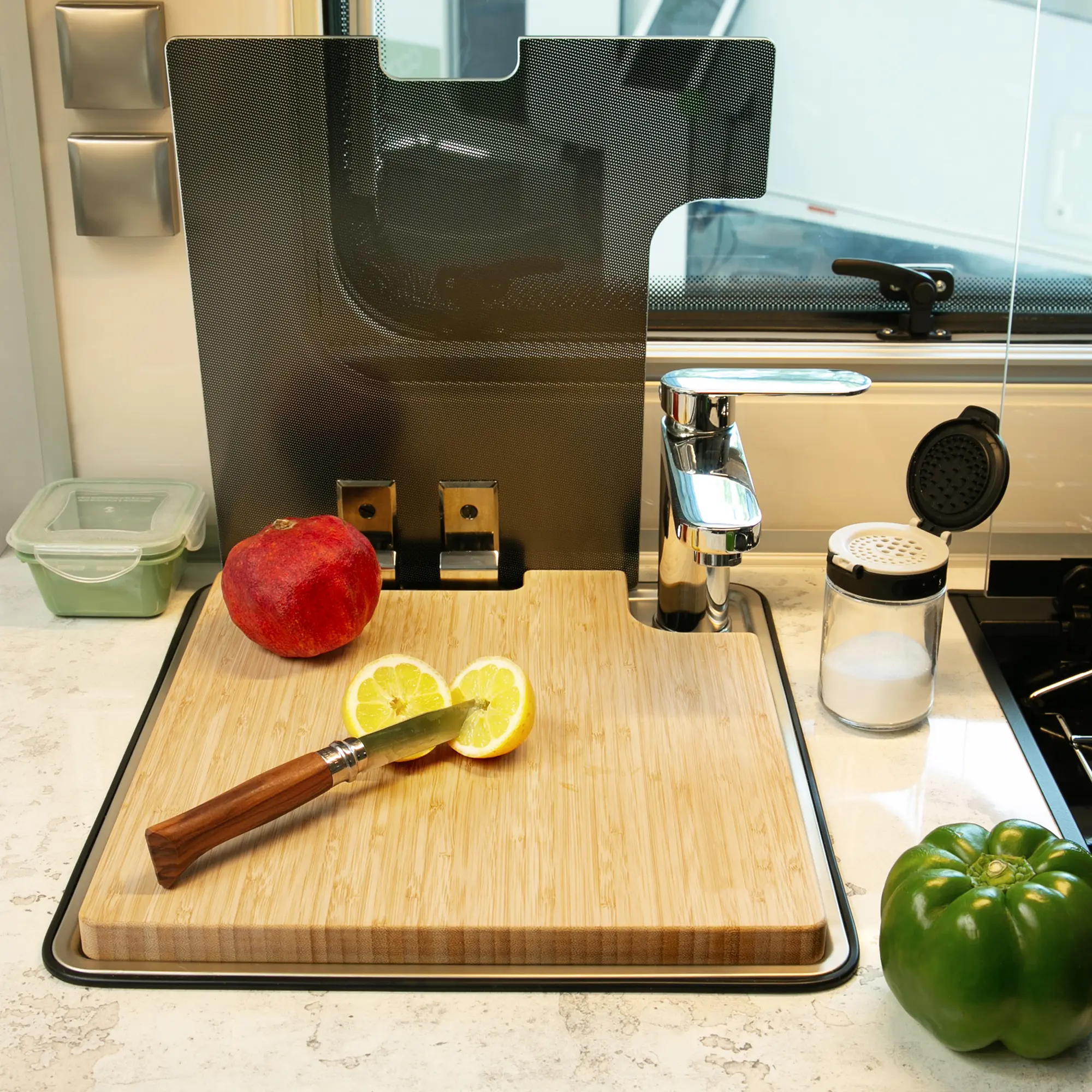 your GEAR Burano bamboo sink cover and cutting board for caravan kitchens, shown fitted over a sink with faucet, featuring a lemon, knife, bell pepper, pomegranate, and modern kitchen accessories.