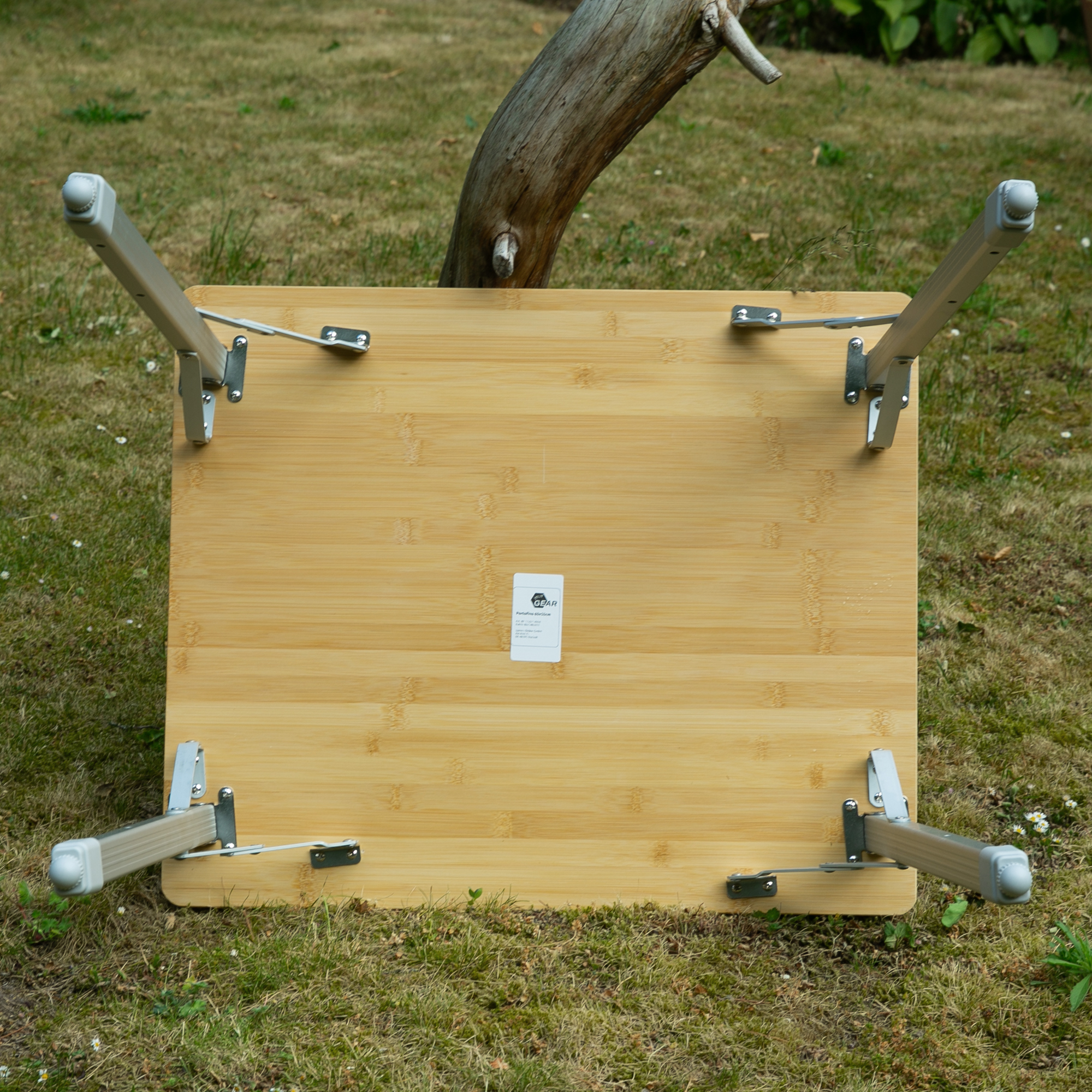 Bambus Campingtisch your GEAR Portofino M mit klappbaren, höhenverstellbaren Aluminiumbeinen, robuste 1,2 cm Bambus-Tischplatte, geeignet für 2 Personen, auf einer Wiese stehend, Unterseite sichtbar.