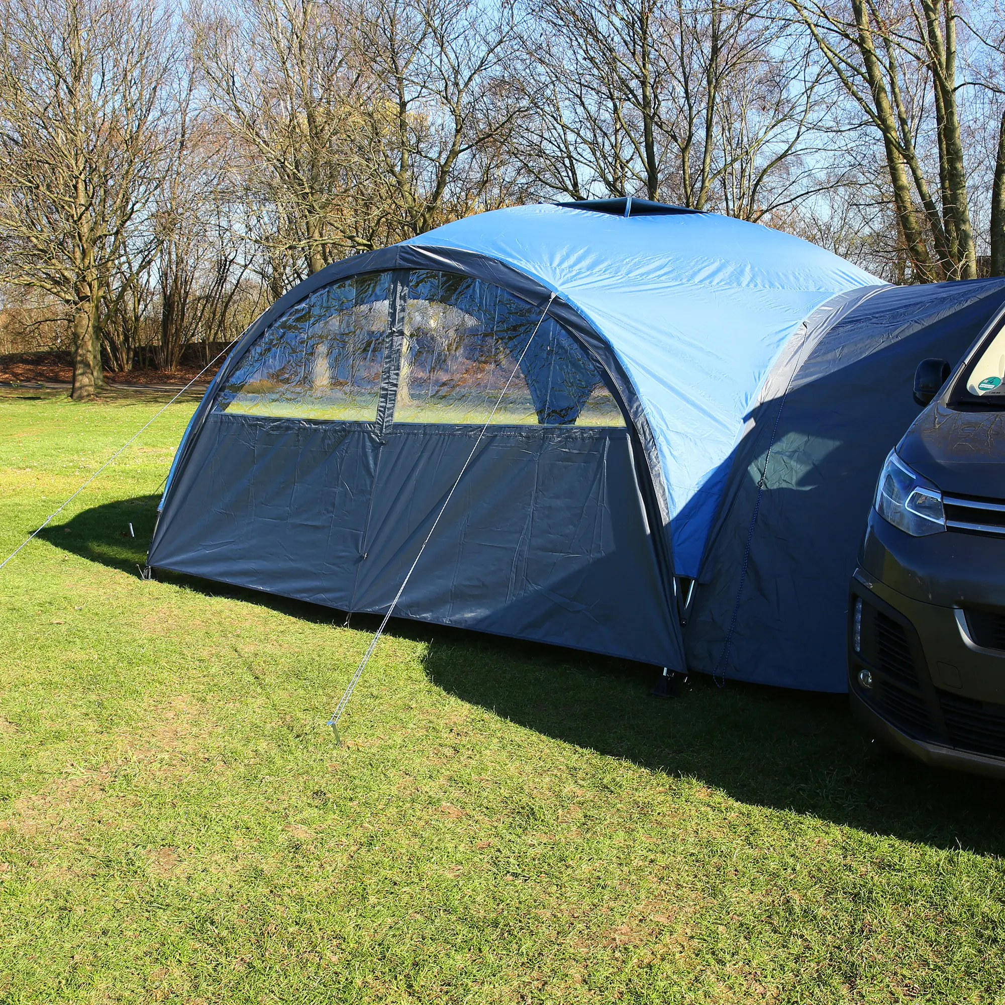 Blue your GEAR Bergamo 430 Bus Connector vehicle awning attached between a camping van and pavilion, with large windows, robust zippers, and tension ropes on a grassy outdoor campsite.