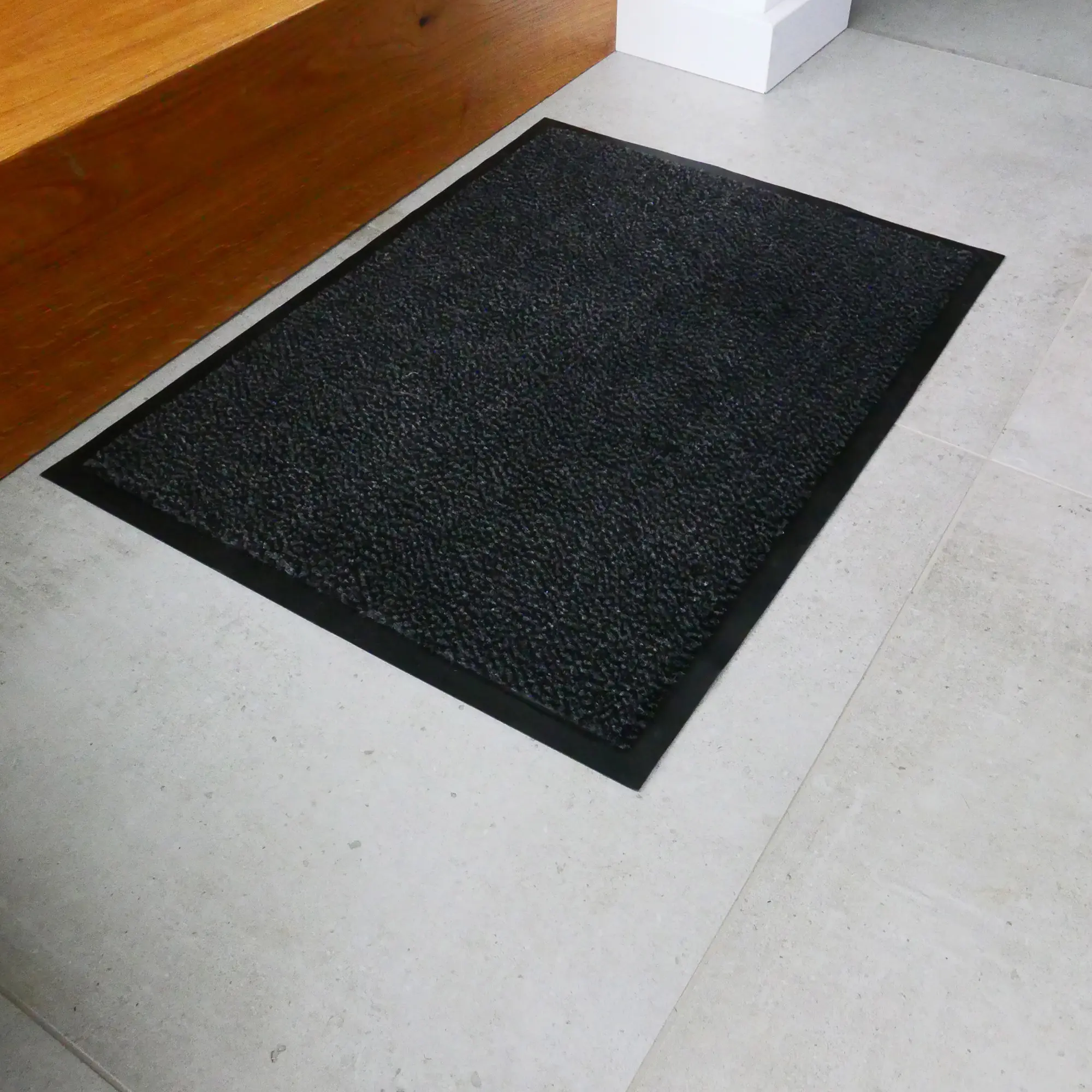 Black yourGEAR dirt-trapping mat 90x60cm with anti-slip rubber edge, dense velour pile, placed on a light tiled floor indoors. Suitable for indoor and outdoor use, washable and UV resistant.
