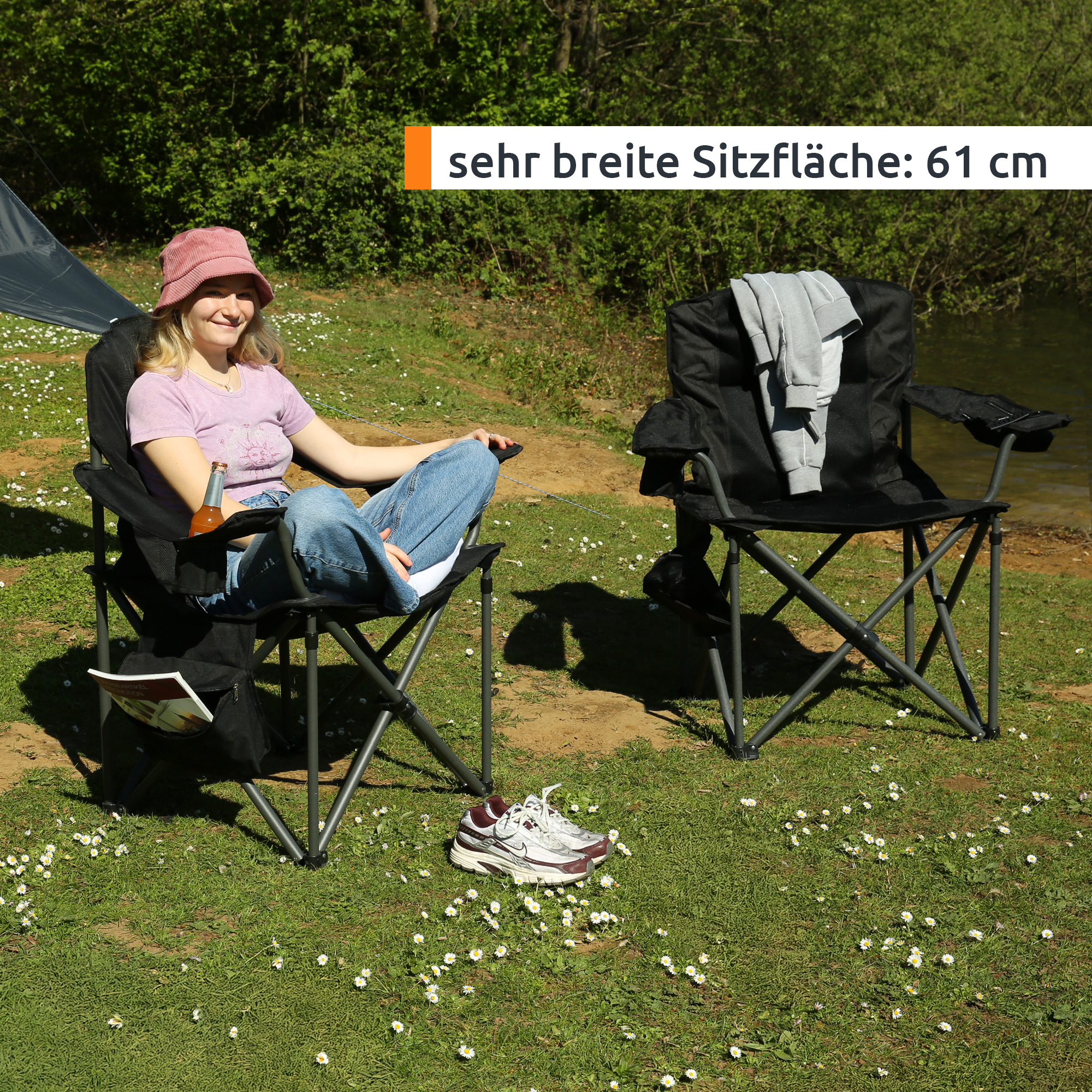 Sedia pieghevole da campeggio nera your GEAR con seduta ampia in 3D‑mesh, robusto telaio tubolare grigio, portabicchiere, tasca organizer laterale e spalliera imbottita, persona seduta all'aperto su prato.