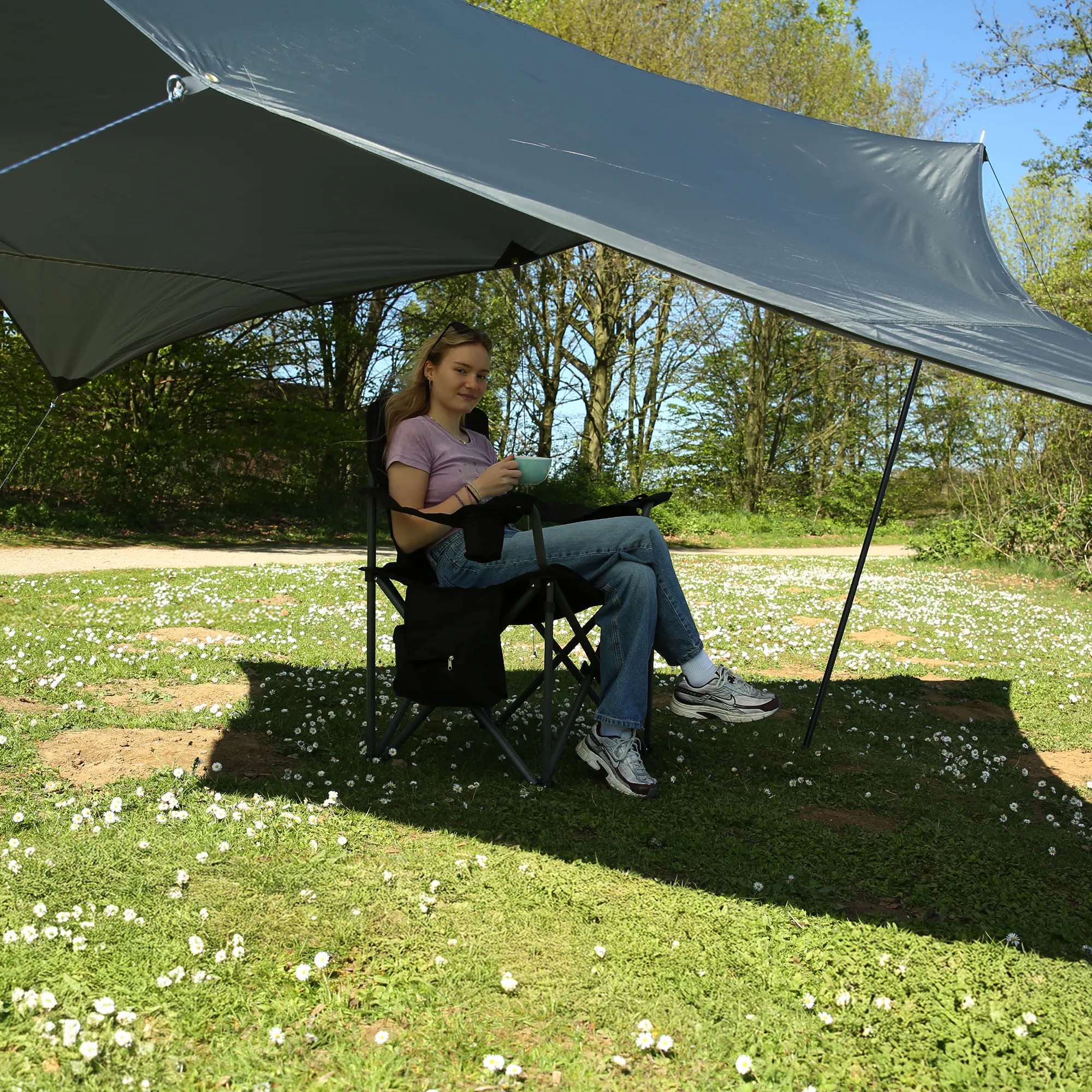 Gray 3x3m your GEAR Atrani tarp sunshade with two black support poles and guy lines, waterproof fabric stretched over a camping chair sheltering a seated woman in a park (UV50+, 5000mm watercolumn)