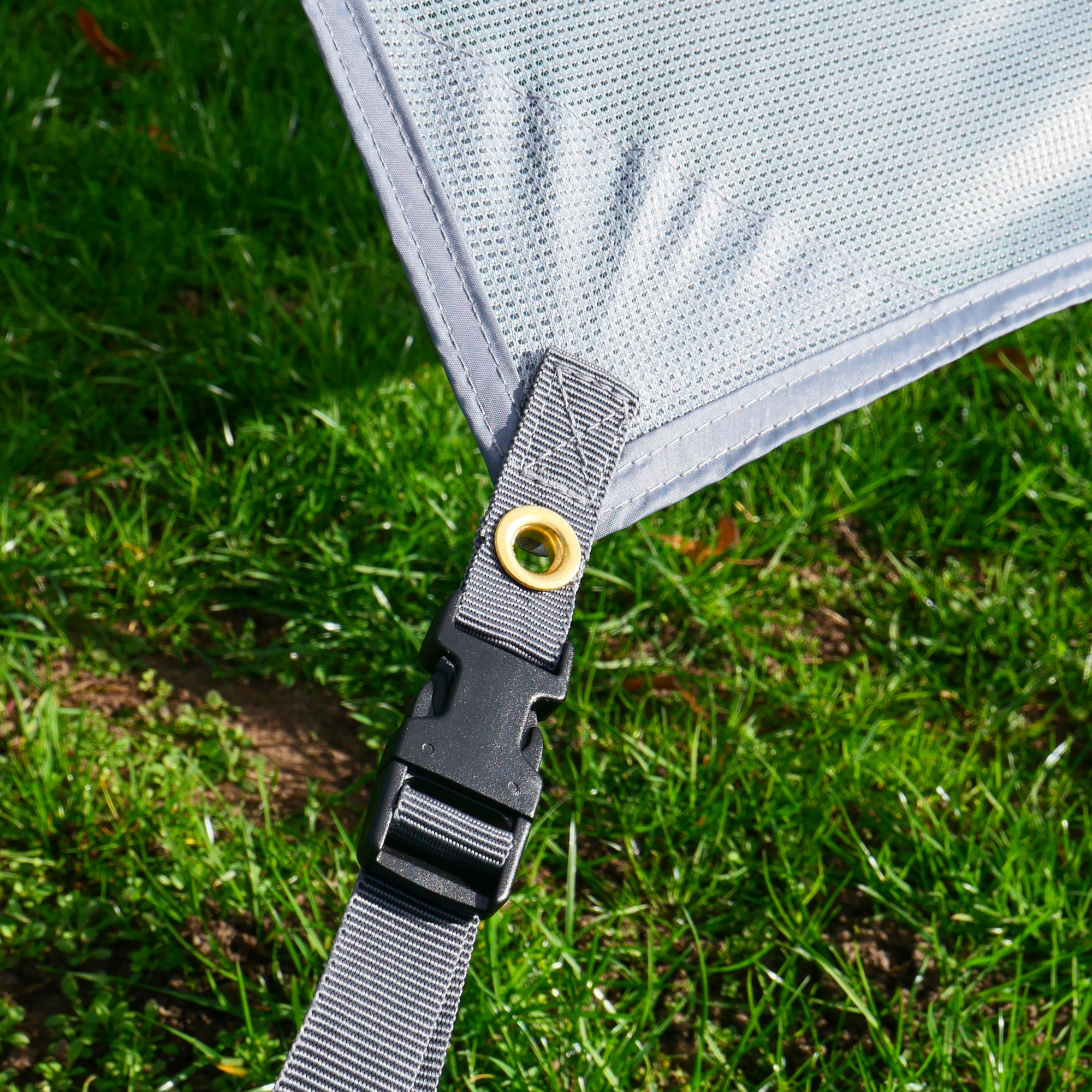 Close-up of the your GEAR Capri Sun Blocker front awning wall, showing grey UV-resistant mesh fabric, reinforced stitching, metal eyelet, and adjustable strap with a black plastic buckle, set on grass.
