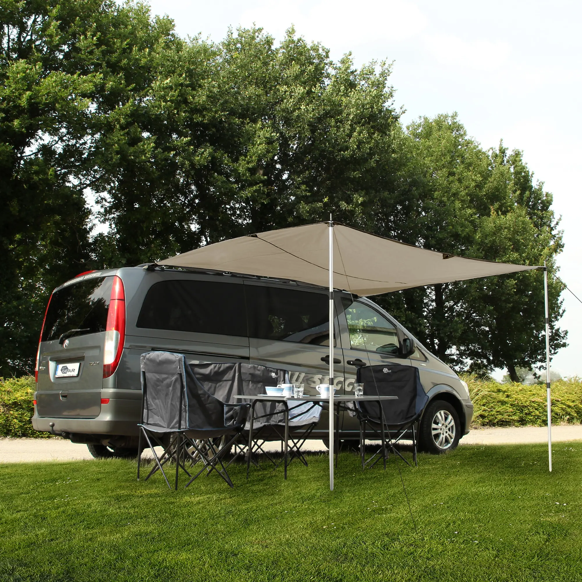 your GEAR Motorhome Cotton Tarp TC 260 x 240 sun canopy attached to a van, set up with two aluminum poles, providing shaded outdoor space with camping table and chairs on grass, ideal for bus, van, or car.