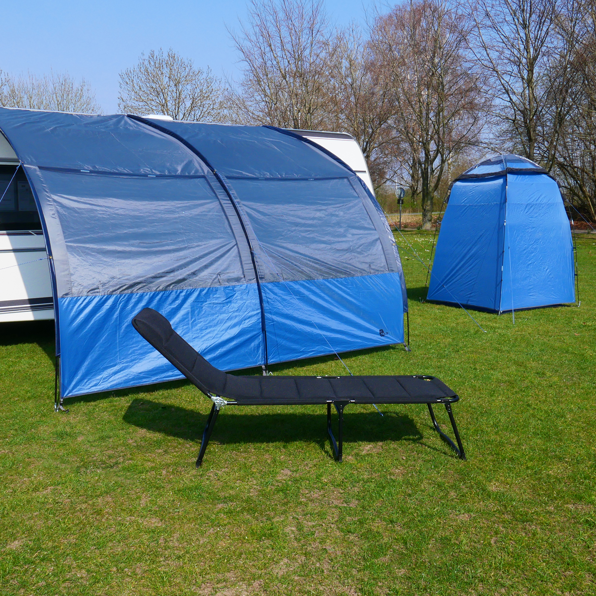 Auvent your GEAR Riviera 350 bleu et gris pour caravane, 3,5x2,6m, avec fenêtres panoramiques, protection UV 50+, monté sur pelouse, abri secondaire bleu et transat noir visibles au premier plan.