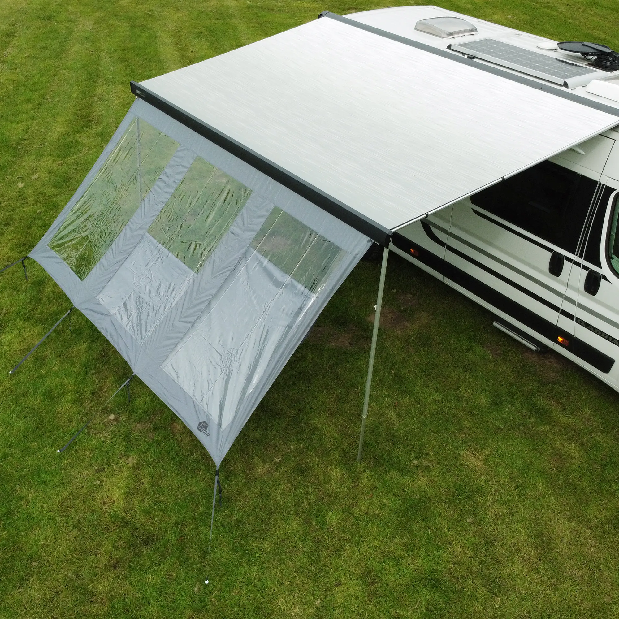 Markisenwand your GEAR Panorama Wall 2,8 x 2 m als Vorderwand an Wohnmobil-Markise montiert, mit großen Klarsichtfenstern, schützt vor Sonne, Wind und Regen, auf grünem Rasen stehend.