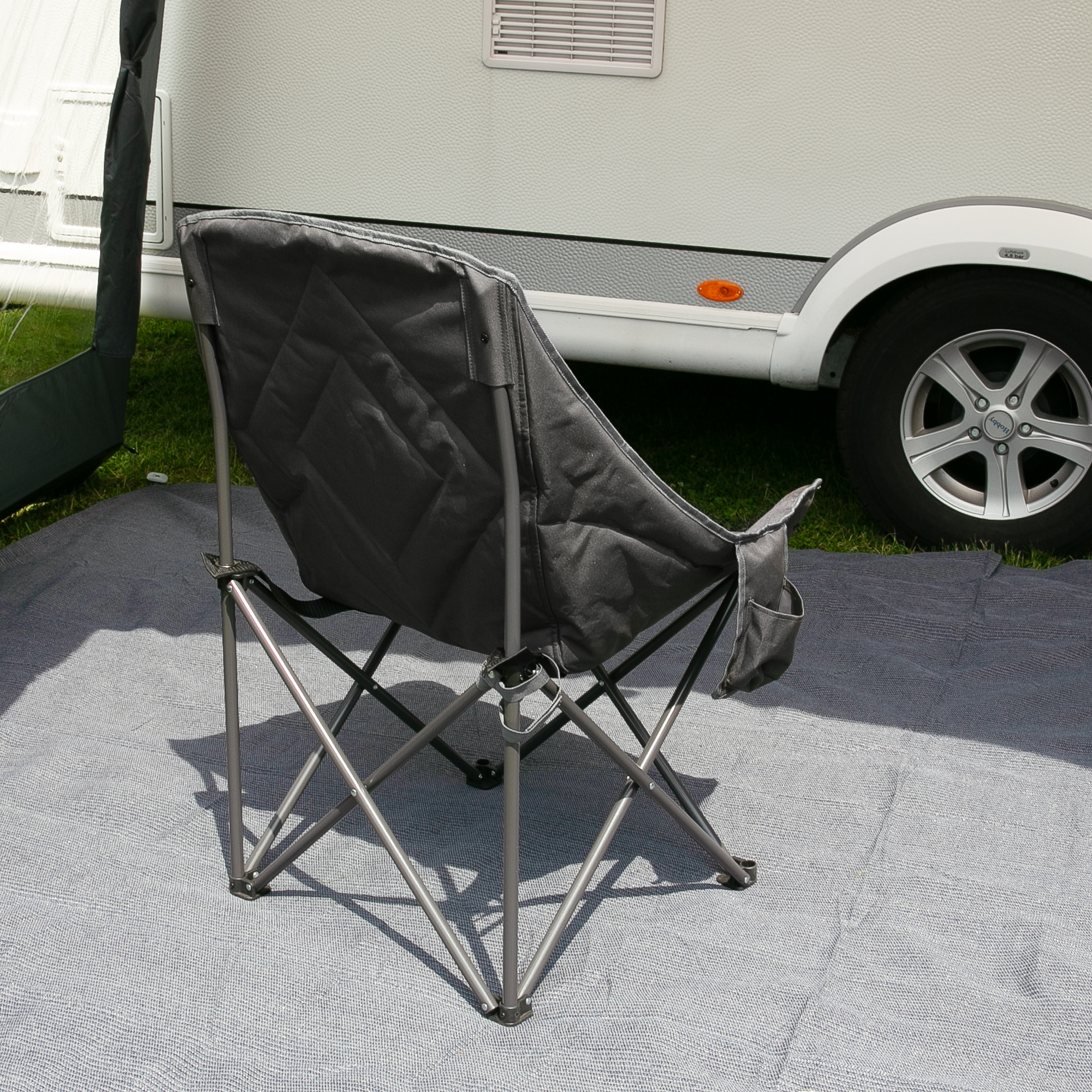 your GEAR Imola XXL camping chair, extra wide with high padded backrest, sturdy steel frame and durable black 600D Oxford fabric, foldable design, shown outdoors next to a caravan on a mat.
