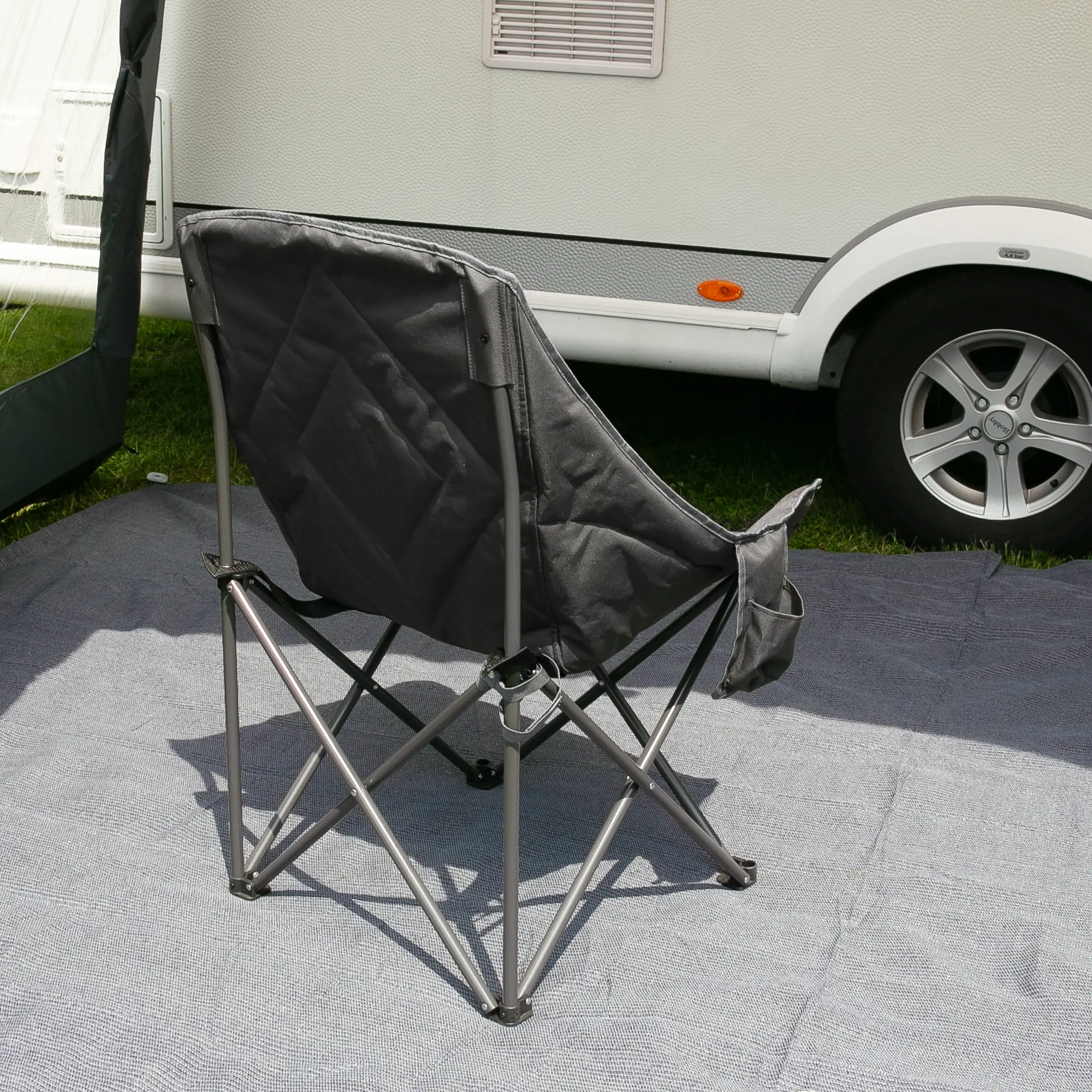 Silla de camping XXL your GEAR Imola, plegable, con respaldo alto y asiento acolchado en tejido negro, estructura de acero resistente y patas anchas, colocada sobre alfombra frente a una caravana.