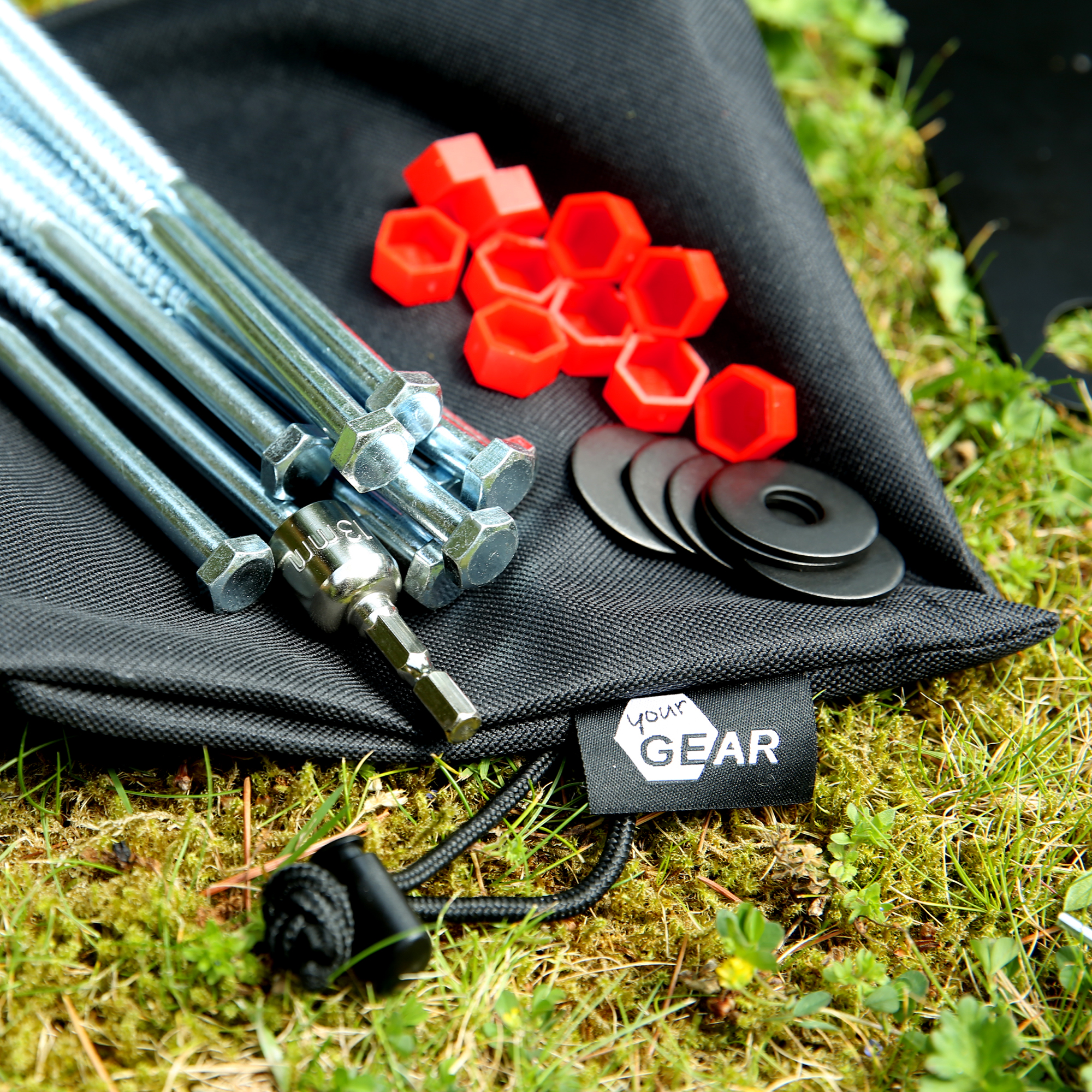 Close-up of your GEAR Montana awning anchor kit: zinc-plated steel bolts and long threaded rods, black rubber washers, red hex-shaped plastic caps and branded black carry pouch on grass, for securing caravan awning feet.