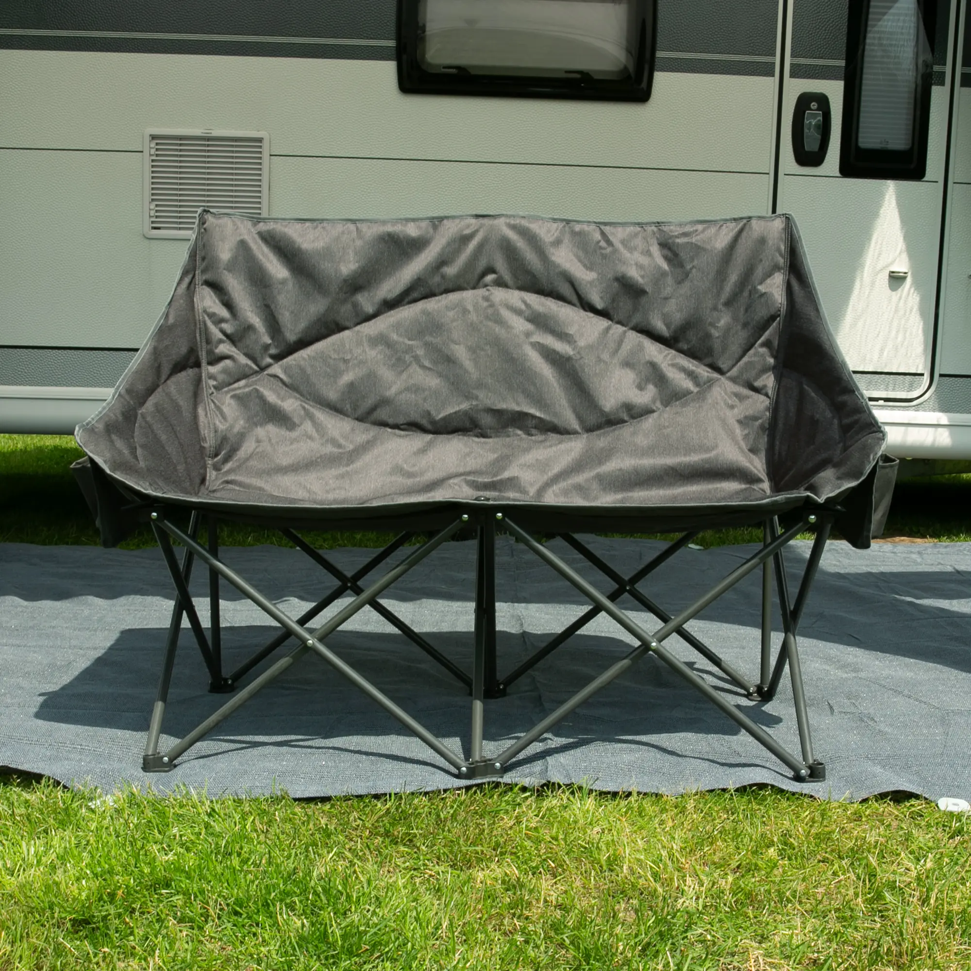 Canapé de camping XXL your GEAR Modena pour 2 personnes, structure en acier, assise et dossier rembourrés, tissu gris two-tone résistant, pieds plats, pliable, installé devant un camping-car sur l’herbe.