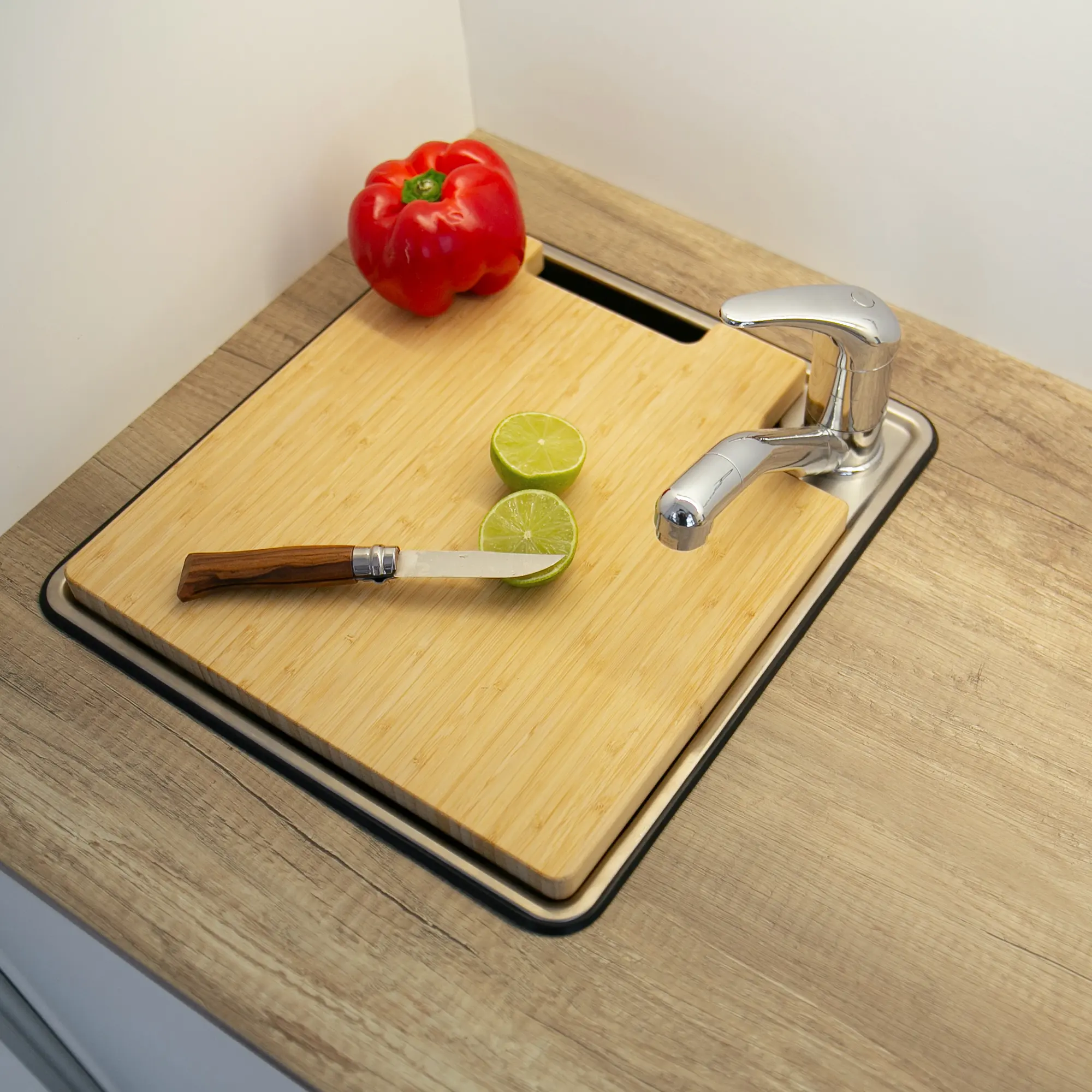 Tabla de cortar y cubierta de fregadero your GEAR Burano de bambú, adaptada para caravanas, sobre fregadero con grifo. Encima hay un cuchillo, una lima partida y un pimiento rojo al fondo.