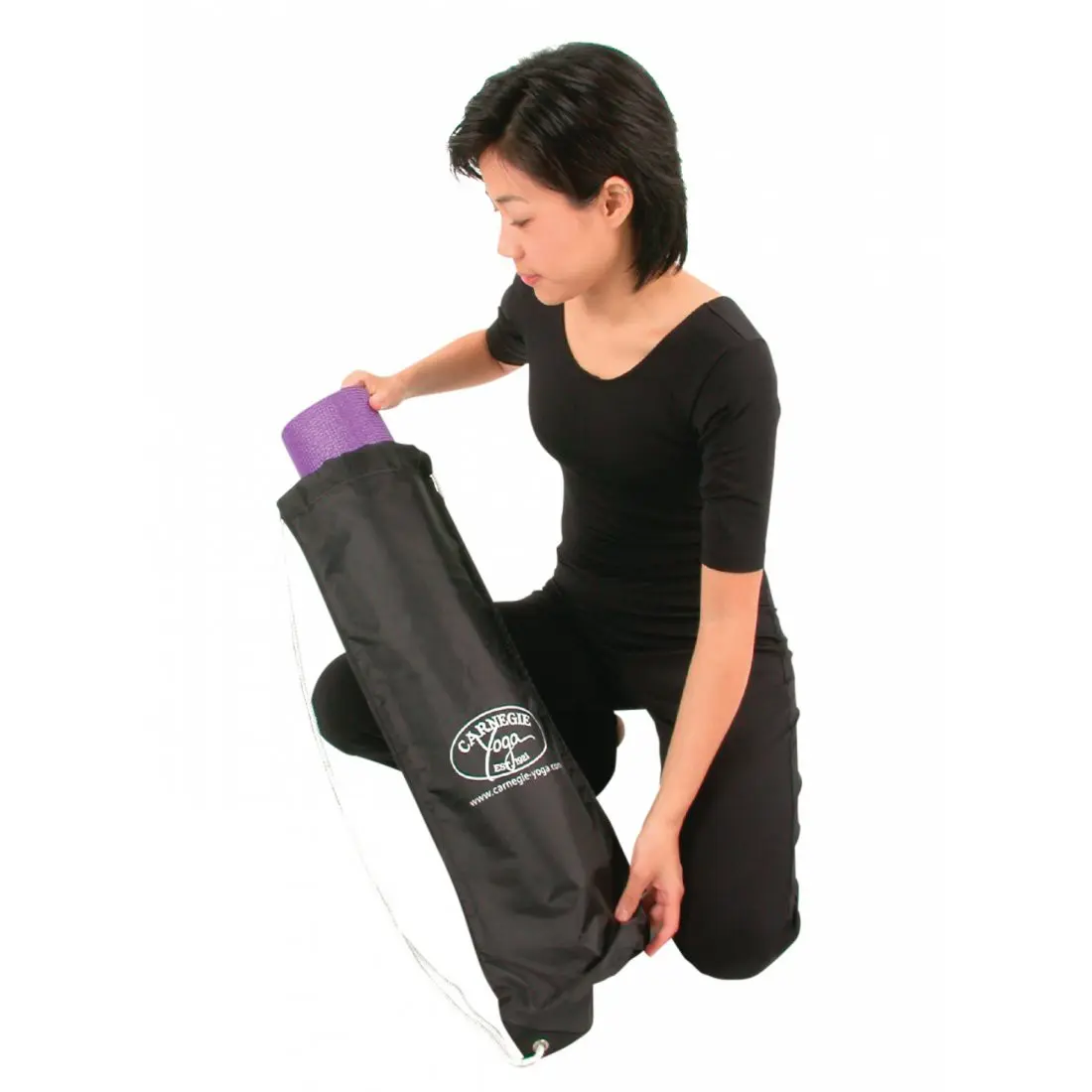 Donna inserisce un tappetino viola arrotolato in una Carnegie Classic Yogatasche nera in nylon, con logo bianco visibile; borsa leggera e impermeabile per tappetini fino a 65 cm x 14 cm di diametro.