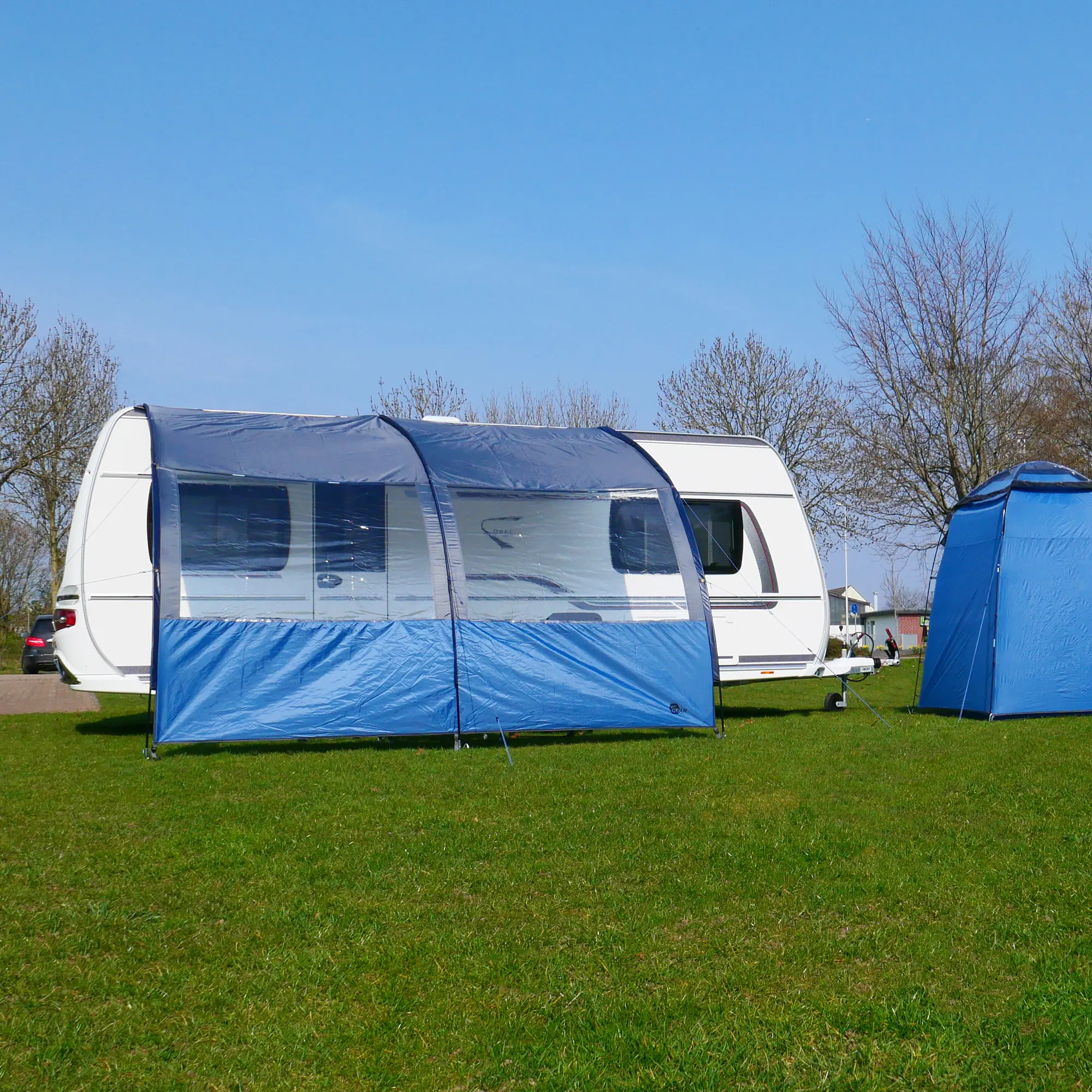 Avance de caravana your GEAR Riviera 200 de 2 x 2,6 m en azul con ventanas panorámicas, protección solar UV 50+ y diseño universal, montado junto a una caravana en un entorno de camping al aire libre.