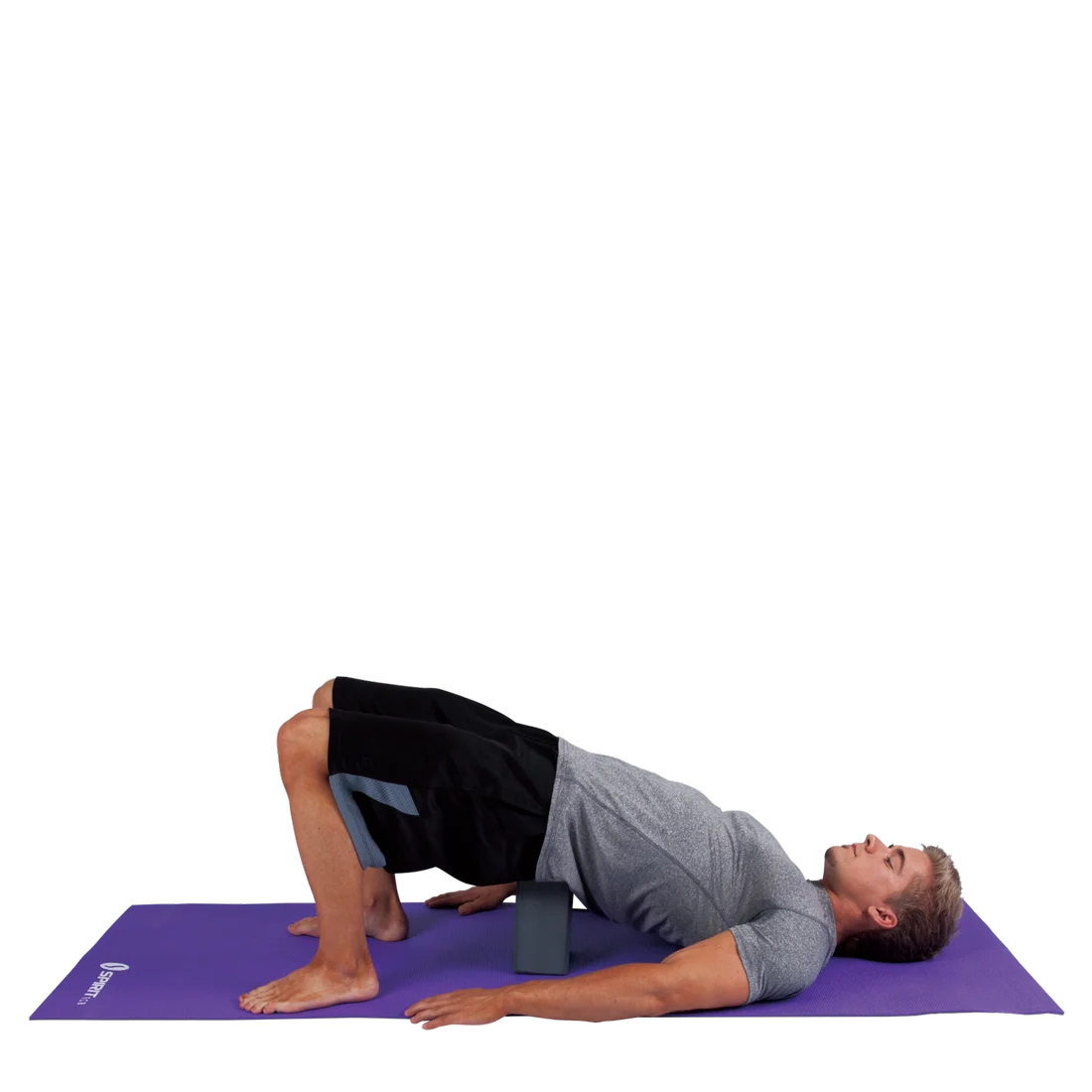 Man practicing yoga on a purple mat using a Spirit Yoga foam block (23x15x10 cm) under the lower back for support; block is dark, rectangular, lightweight, and designed for stability during poses.