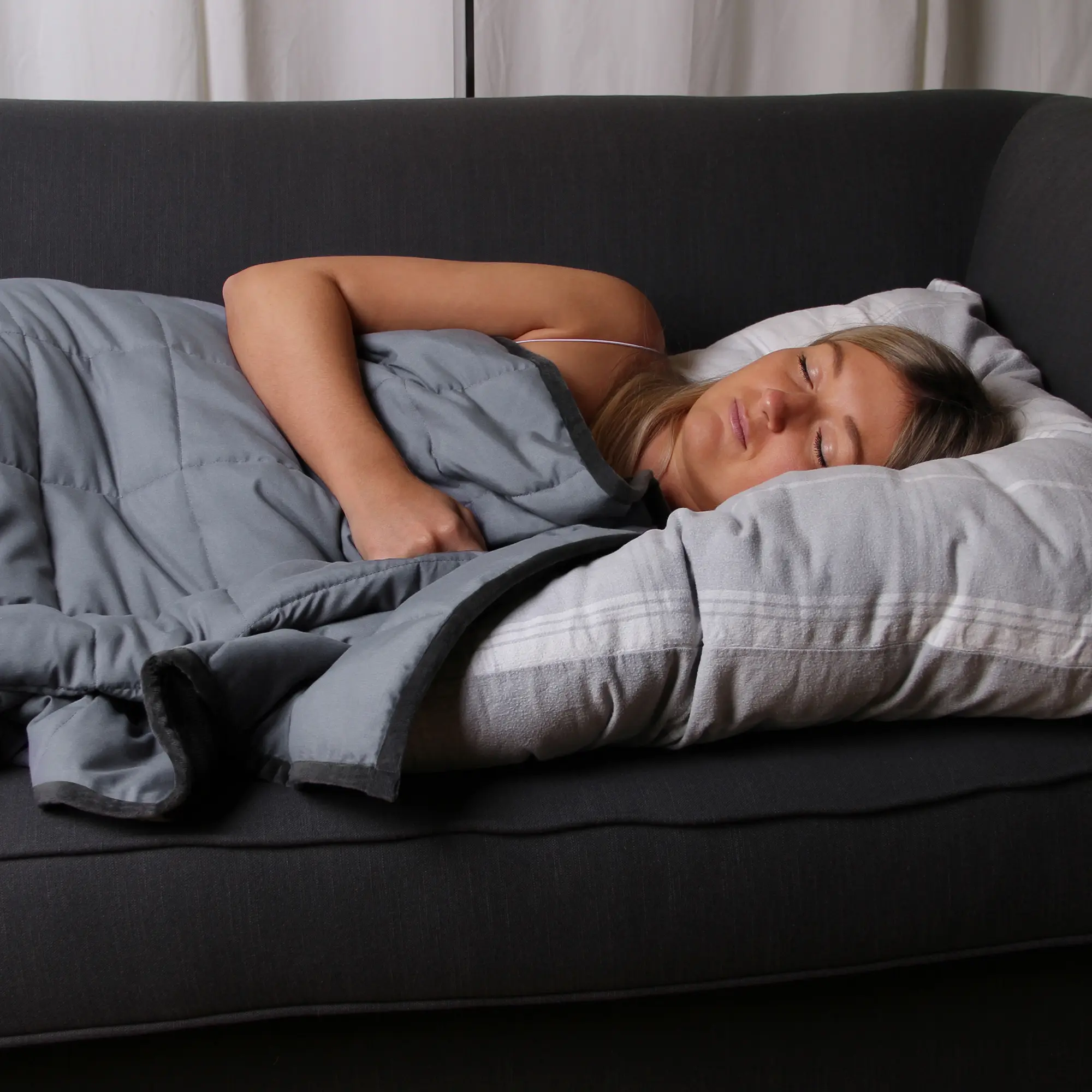 Donna che dorme coperta da una yourGEAR Como coperta terapeutica ponderata da 6 kg in cotone grigio, dimensioni 135x200 cm, su un divano scuro, ambiente rilassante per il riposo e la calma.