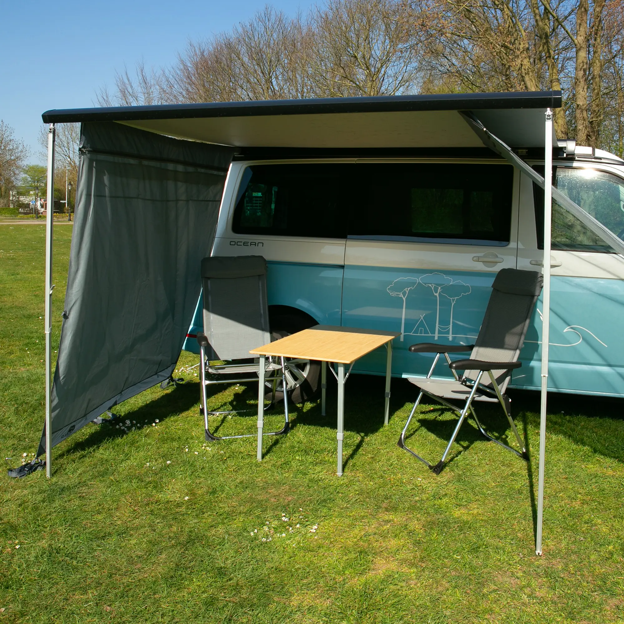 Bambus Campingtisch your GEAR Portofino L für 4 Personen, klappbar, höhenverstellbar mit Teleskopbeinen, wetterfest, neben Campingbus mit zwei Stühlen auf einer Wiese aufgestellt.