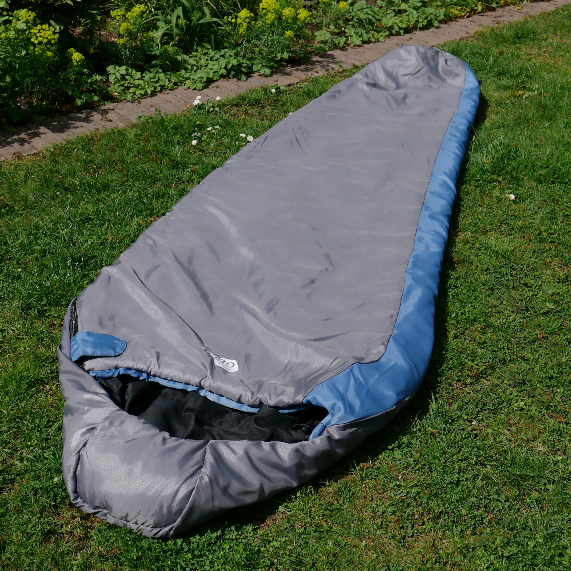 yourGEAR Schlafsack Pali -3°C, Mumienschlafsack in Grau mit blauen Akzenten, 220x75cm, leicht, kompaktes Packmaß, ideal für Sommer, Camping und Outdoor, auf grünem Rasen ausgebreitet.