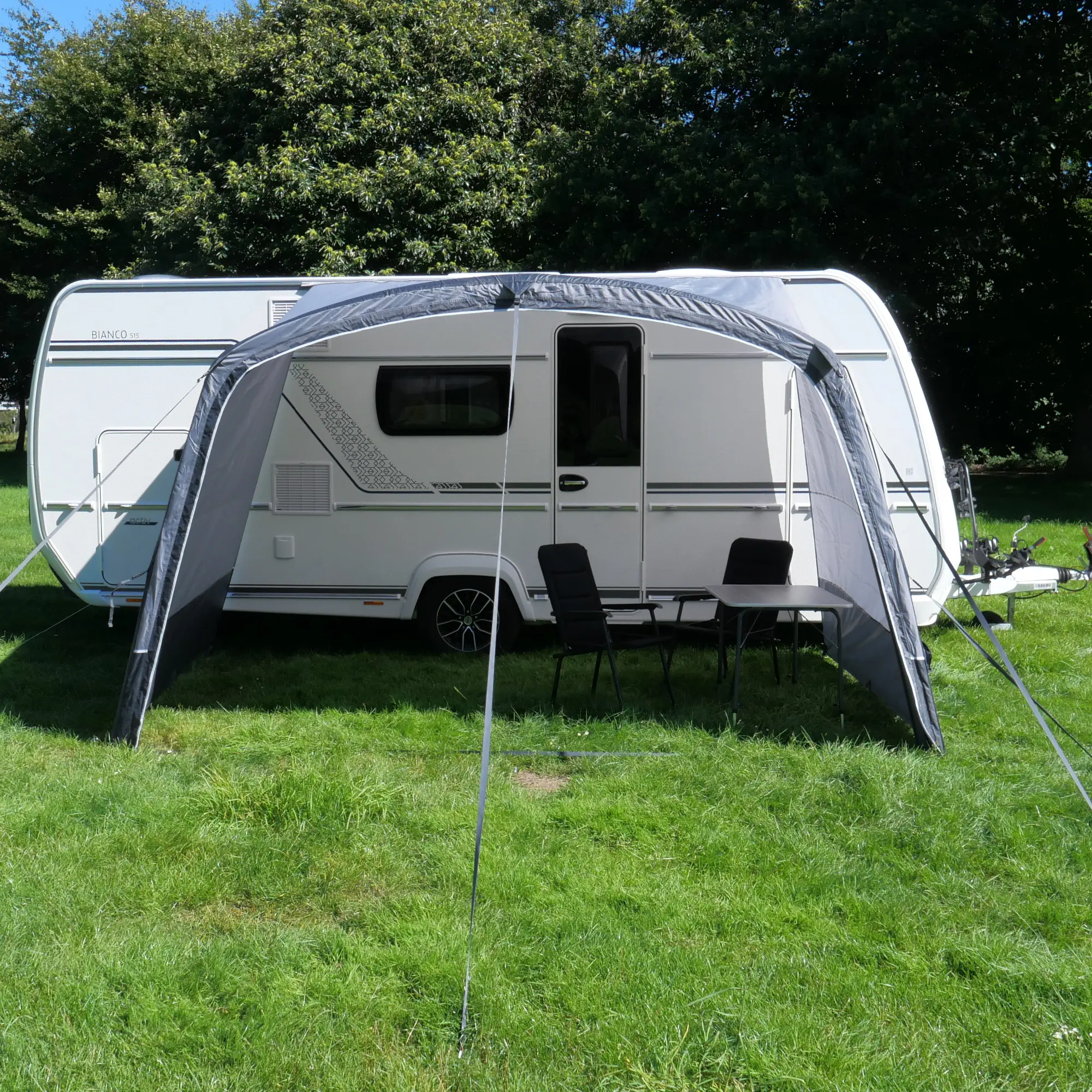 Aufblasbares your GEAR Otranto Air Sonnendach 380 x 250 cm am Wohnwagen, mit Panoramawand, schwarzem Gestänge, befestigt auf Rasen, darunter Tisch und zwei Stühle, idealer Sonnenschutz für Camping.