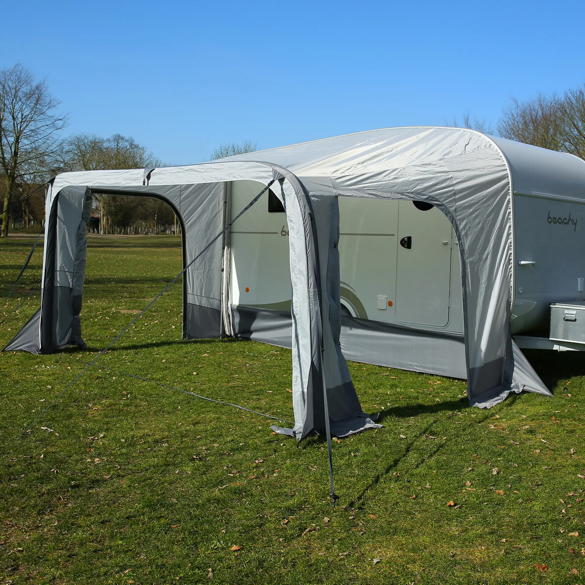Auvent gris installé devant une caravane blanche sur une pelouse, sous un ciel bleu et dégagé, dans un environnement naturel avec quelques arbres en arrière-plan.