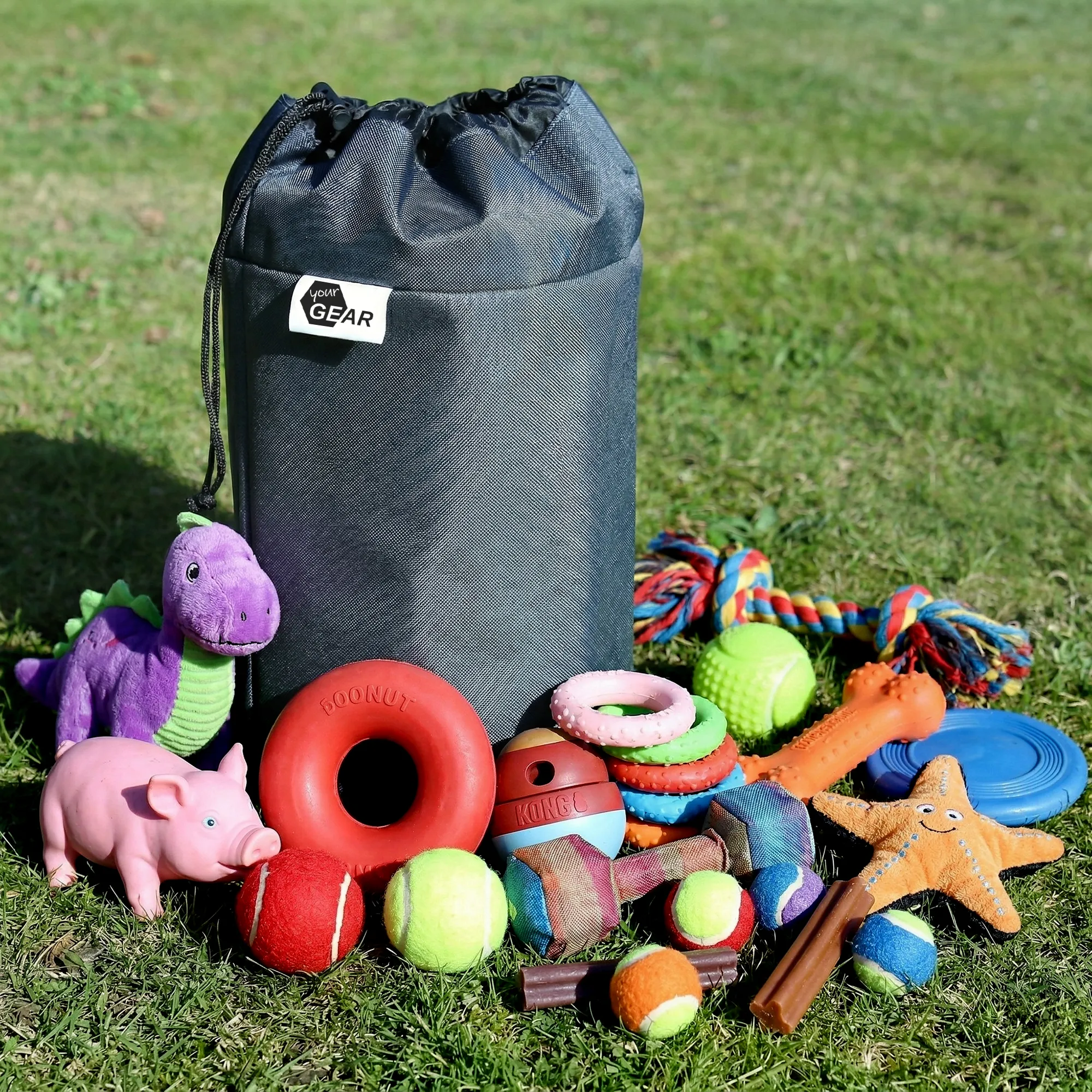 Grand sac gris pour jouets posé sur l'herbe entouré de peluches et nombreux jouets colorés pour chien : balles, anneaux, corde, hochet et figurines.