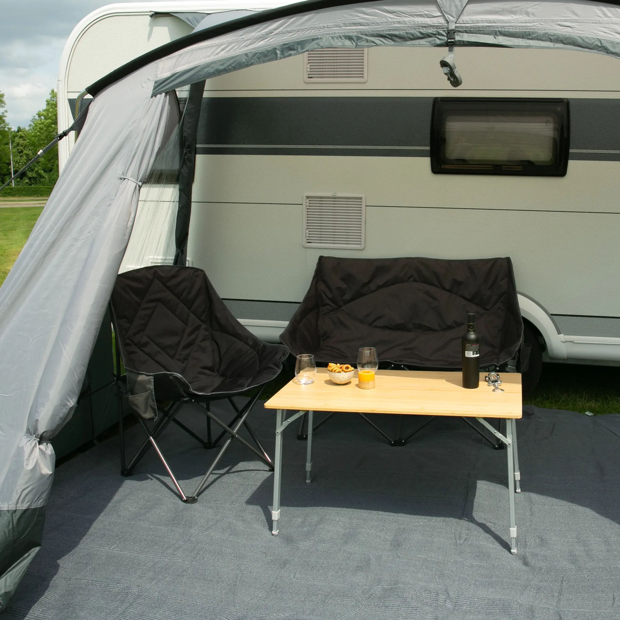 Table de camping your GEAR Portofino L en bambou pour 4 personnes, plateau rectangulaire naturel, pieds télescopiques réglables en hauteur, installé devant une caravane avec chaises pliantes noires et boissons dessus.