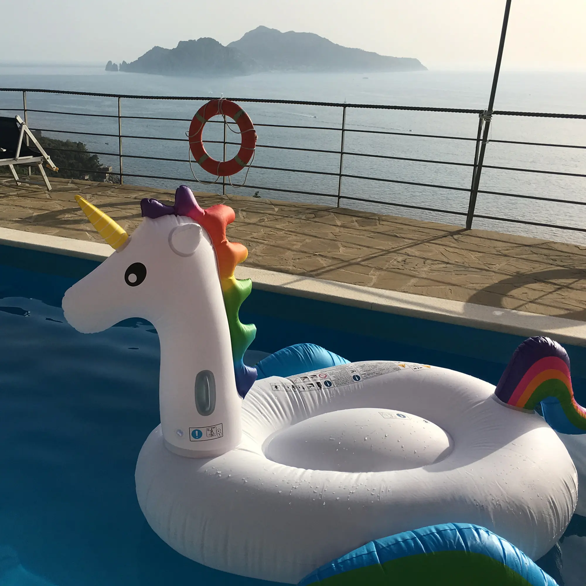 Unicorno gonfiabile XXL Jilong 220x160x120 cm, bianco con criniera e coda arcobaleno, due maniglie ergonomiche, galleggiante per piscina adatto ad adulti e bambini, vista mare sullo sfondo.