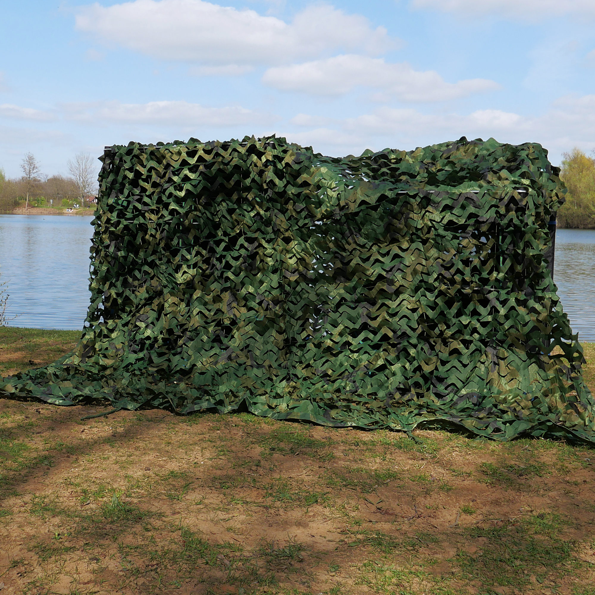 Red de camuflaje yourGEAR de 1,5 x 5 m, patrón militar verde, instalada al aire libre junto a un lago, ideal para sombra, ocultación y protección solar en jardín, caza o actividades exteriores.