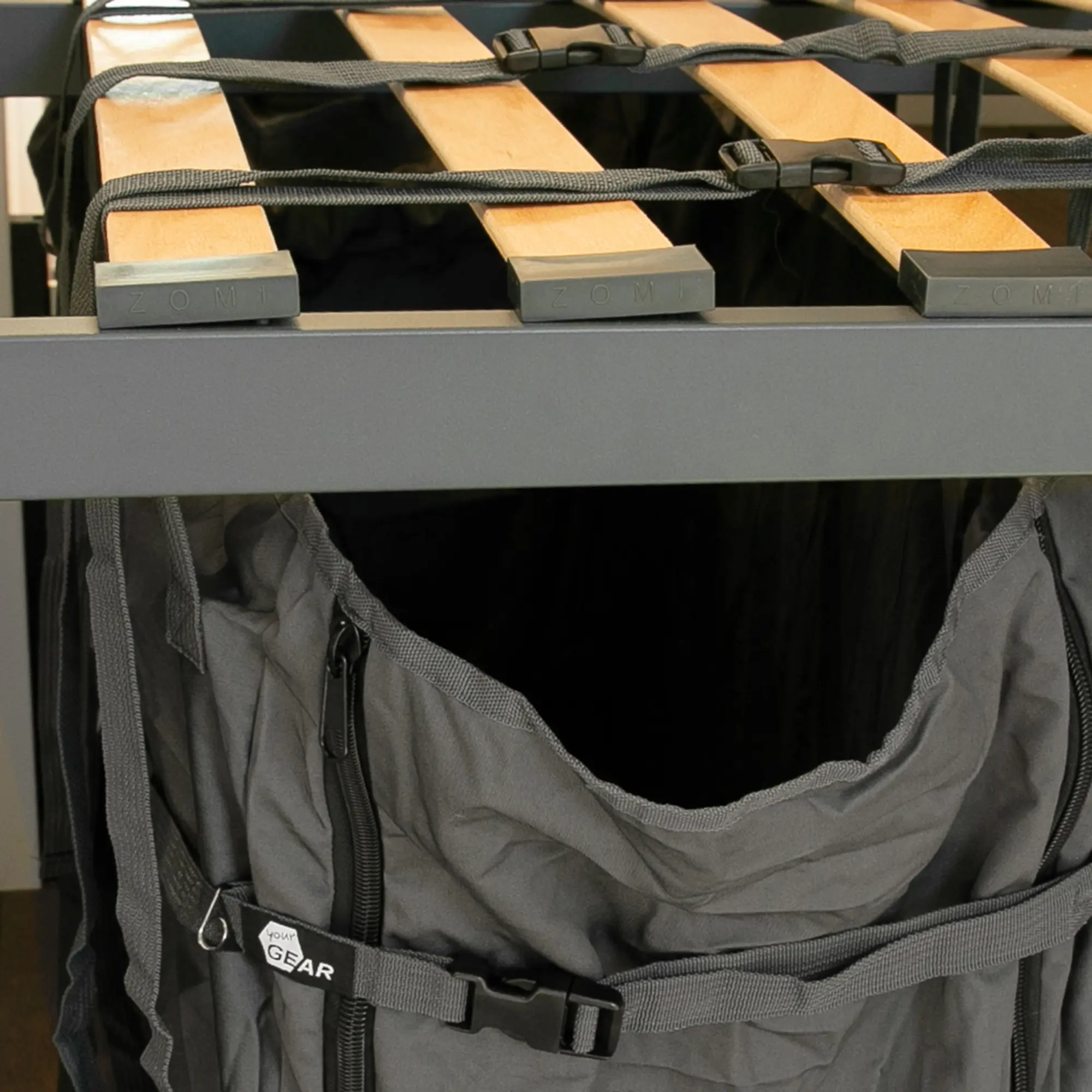 your GEAR Stauraumtasche Bozen für Campingmöbel, vertikal montiert in der Camper-Van Heckgarage, mit Reißverschluss, Gurten und robustem Stoff, unter einem Holzlattenrost befestigt.