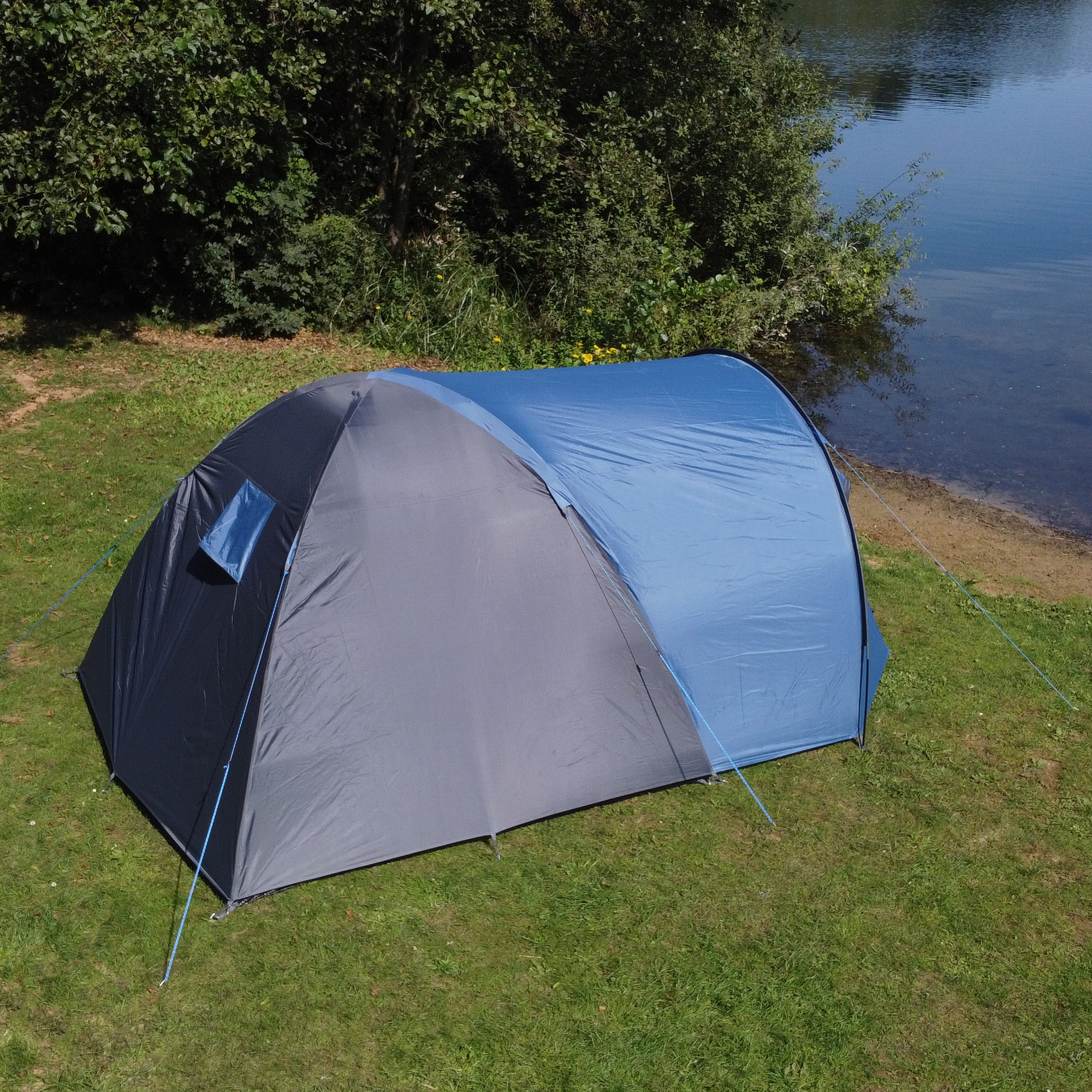 your GEAR Cetona 4-person dome tent in blue and grey, with large vestibule, side entrance, and standing height, set up on grass near water, ideal for family camping or festivals.